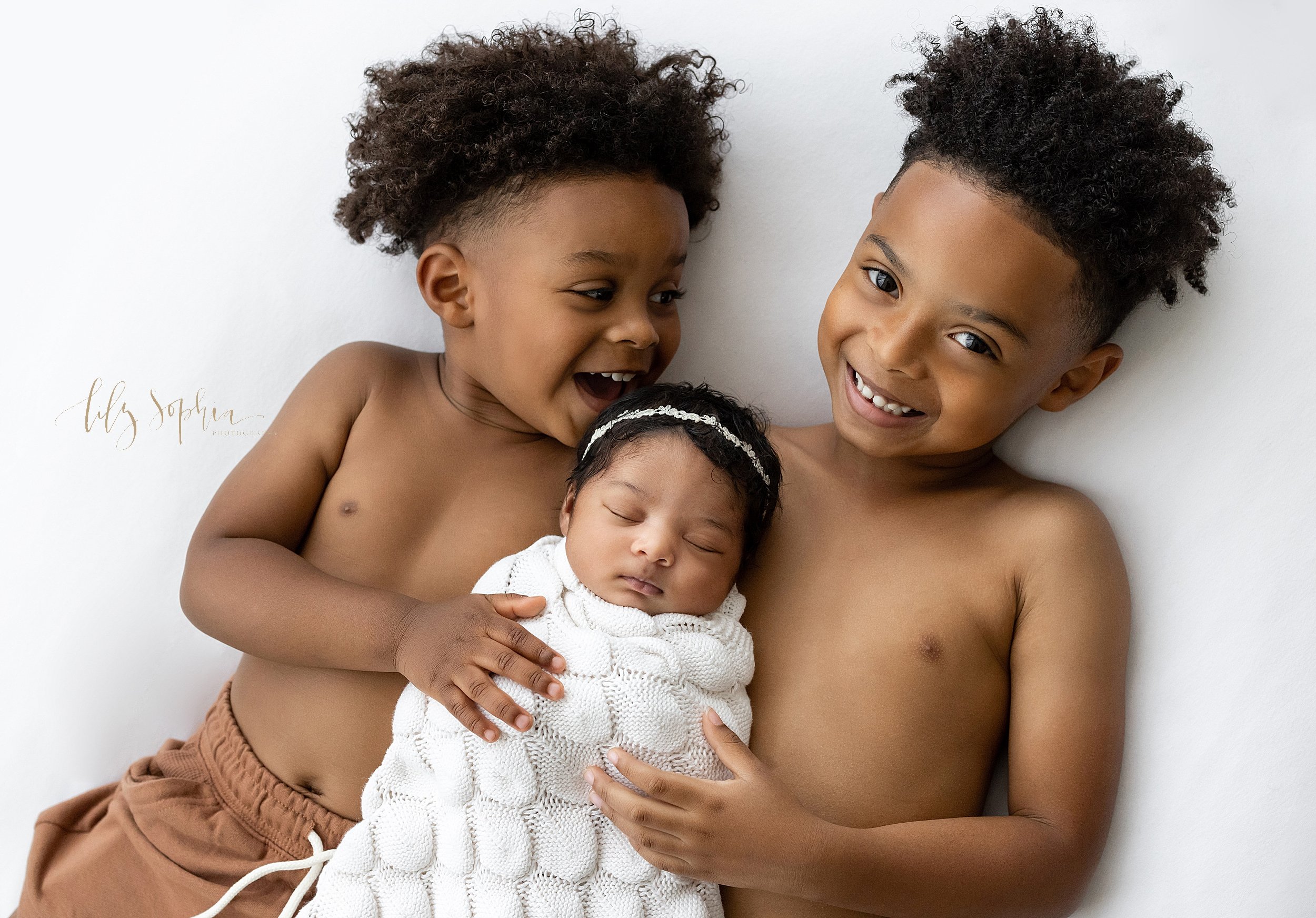  Family newborn portrait of brothers lying on their backs on a bed with their  sleeping newborn baby sister swaddled to her chin with a soft white knitted blanket lying between them taken in a studio near Decatur in Atlanta that uses natural lighting