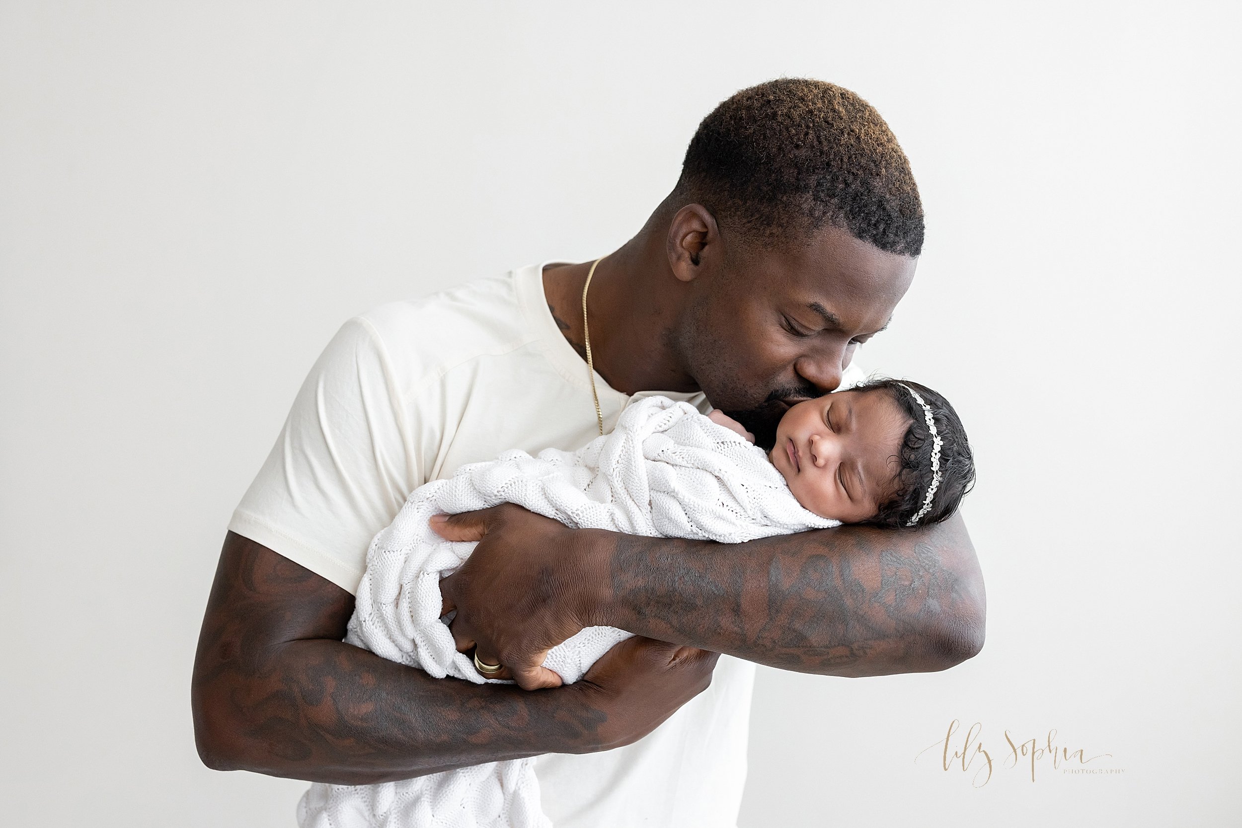  Newborn portrait of an African-American father cradling his newborn daughter in his arms as he kisses her cheek taken using natural light in a studio near Sandy Springs in Atlanta. 