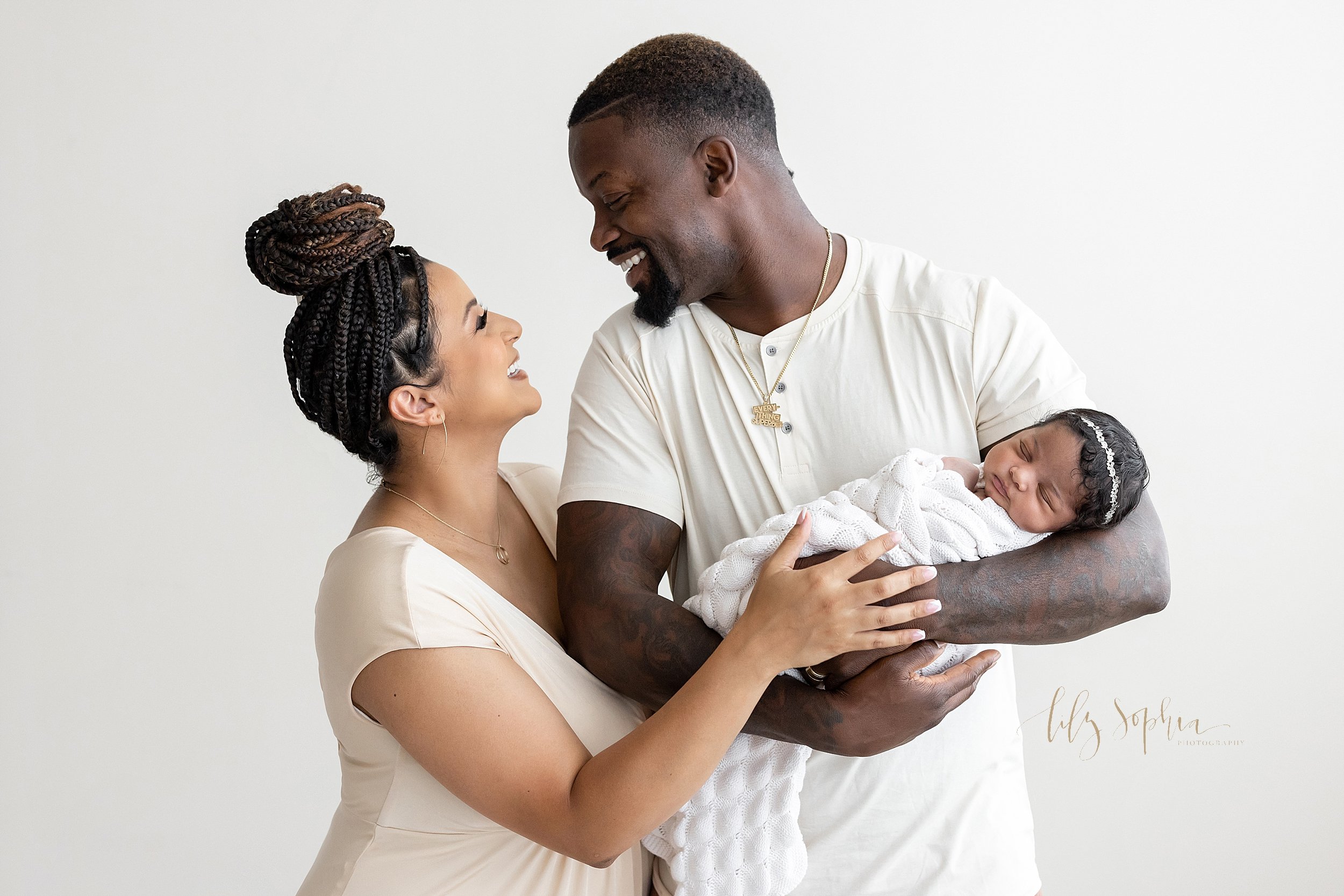  Newborn family photo of an African-American father cradling his newborn daughter in his arms as his wife stands next to him and they look into one another’s eyes with pride and joy taken in front of a window streaming natural light in a studio near 