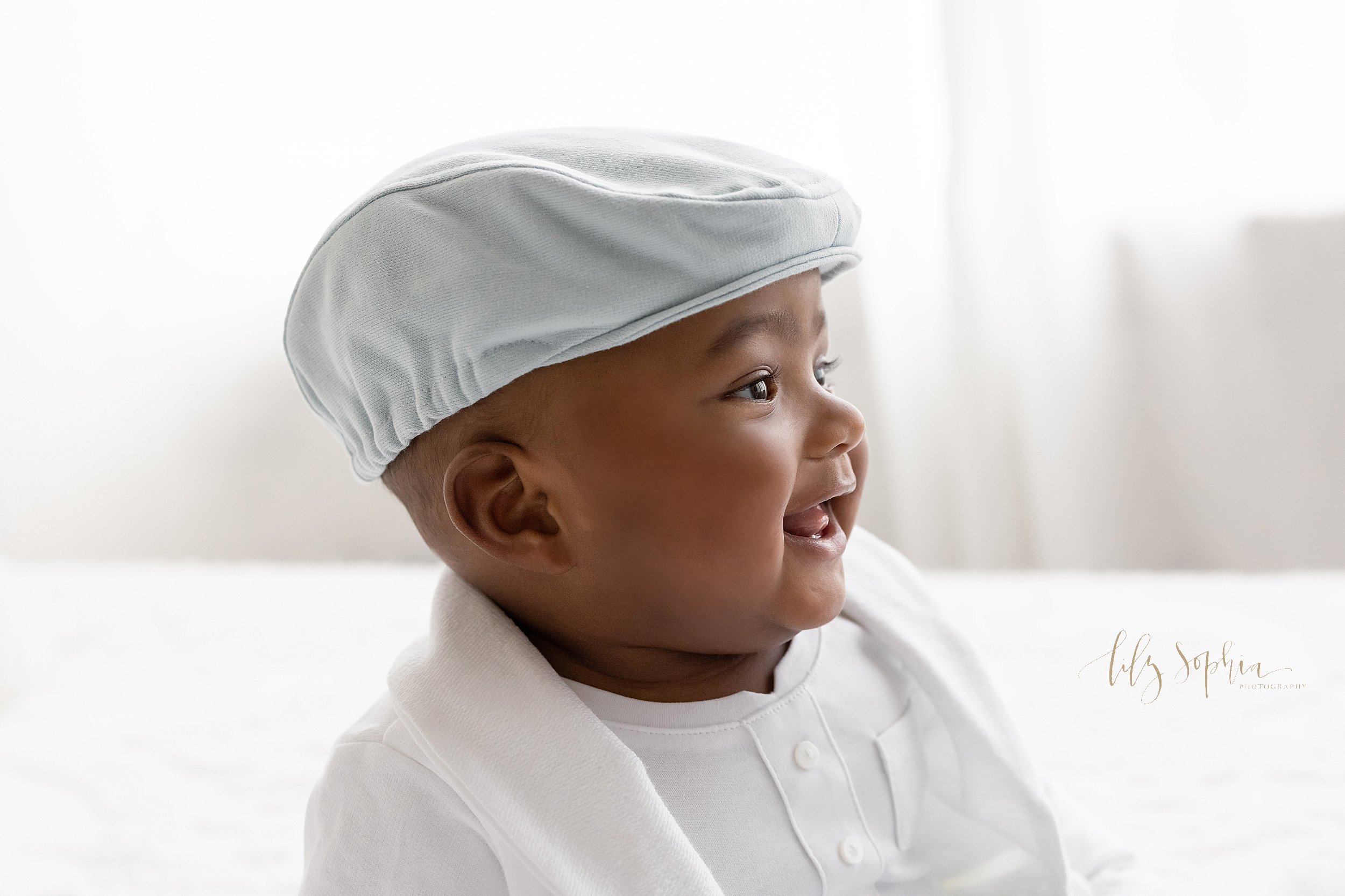  Milestone baby portrait of an African-American baby boy sitting on a bed wearing a white derby cap, a white suit jacket, and a white collarless shirt taken in a natural light studio near Midtown in Atlanta. 