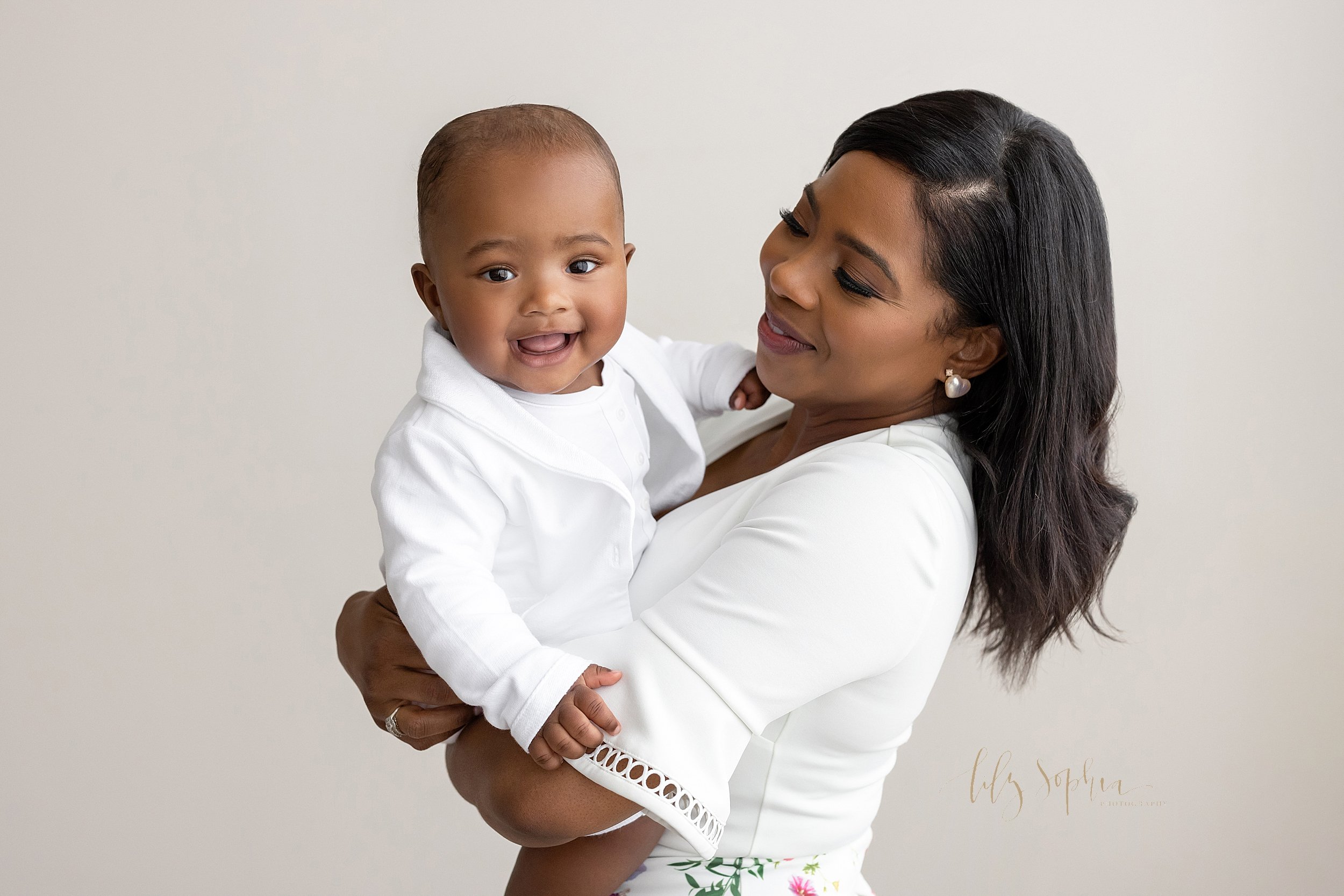  Family photo session of an African-American mother dressed in a white short bell sleeved shirt holding her baby boy dressed in a white collarless shirt and a white suit jacket in her arms taken near Decatur in Atlanta in a natural light studio. 