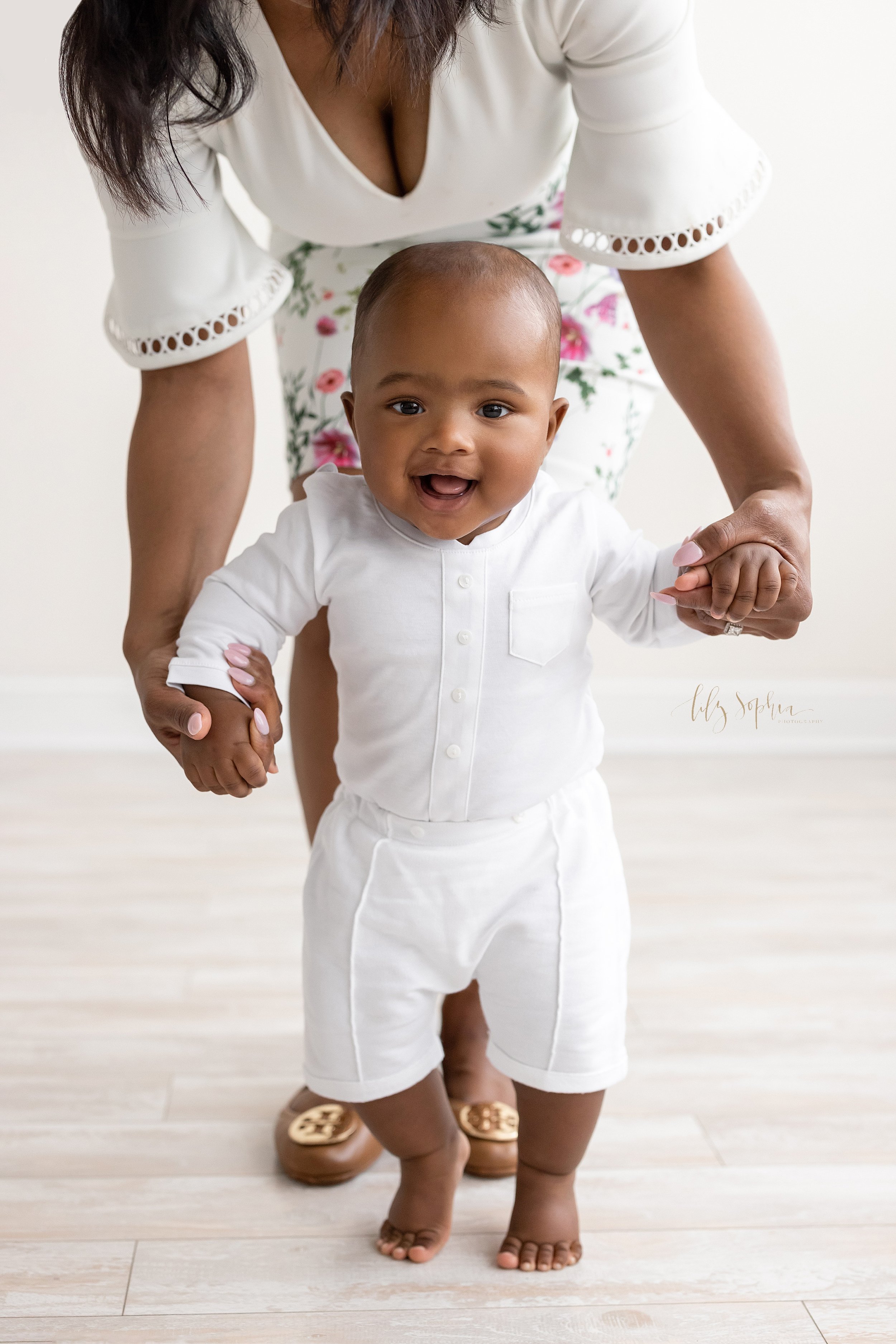  Milestone baby photo of an African-American baby boy with his mom holding his hands as she helps him walk in front of her taken in front of a window streaming natural light in a studio near Poncey Highlands in Atlanta, Georgia. 