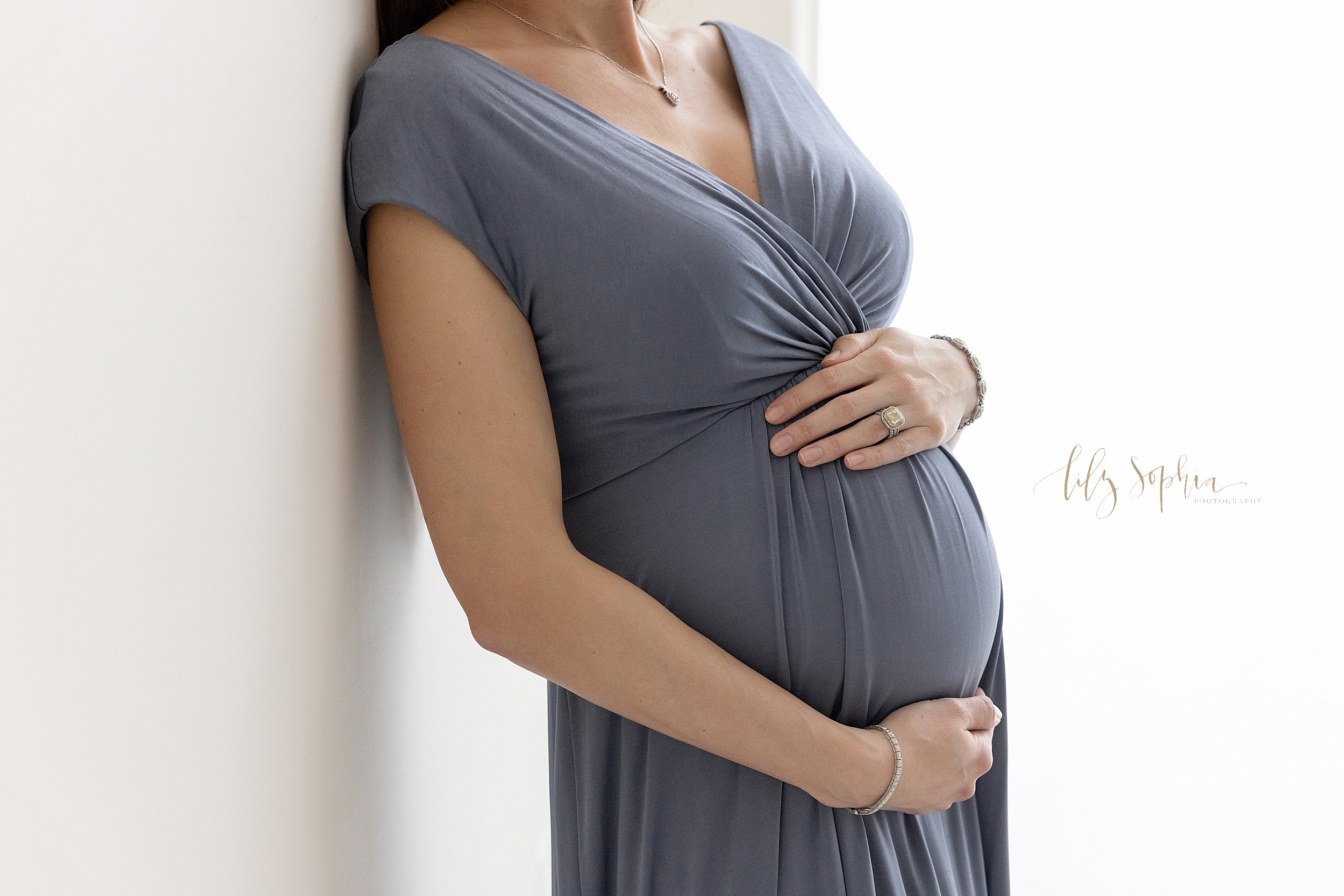  Close-up maternity photo of the belly of a pregnant woman as she stands in a blue knit gown with her back against a wall as natural light streams in a window next to her and she frames her belly with her hands taken in a studio near Alpharetta in At