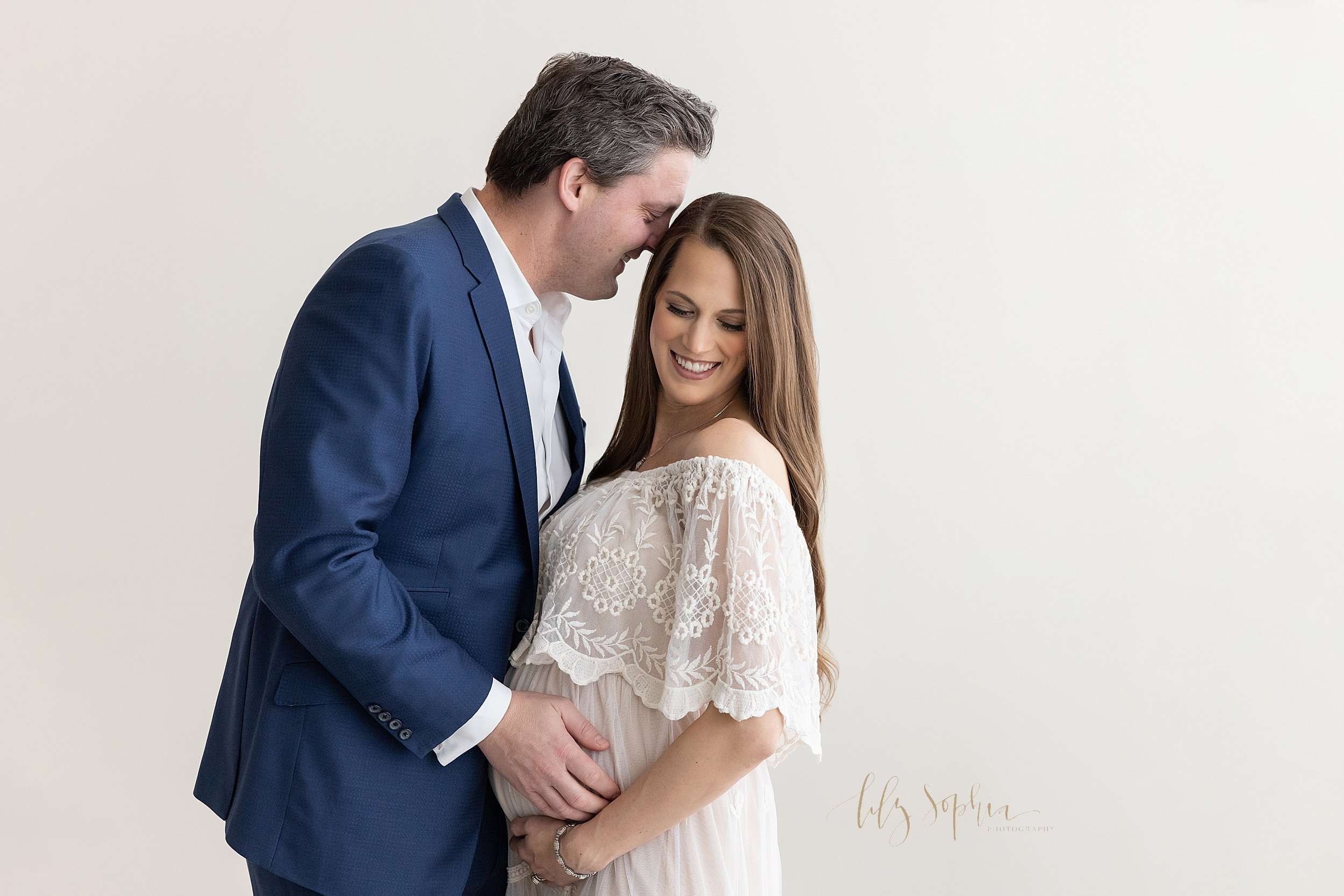  Maternity photo session of a father facing his wife as he places his right hand on his child in utero and whispers in his wife’s ear while she turns her head over her left shoulder and holds the base of her belly as the couple stands in a natural li