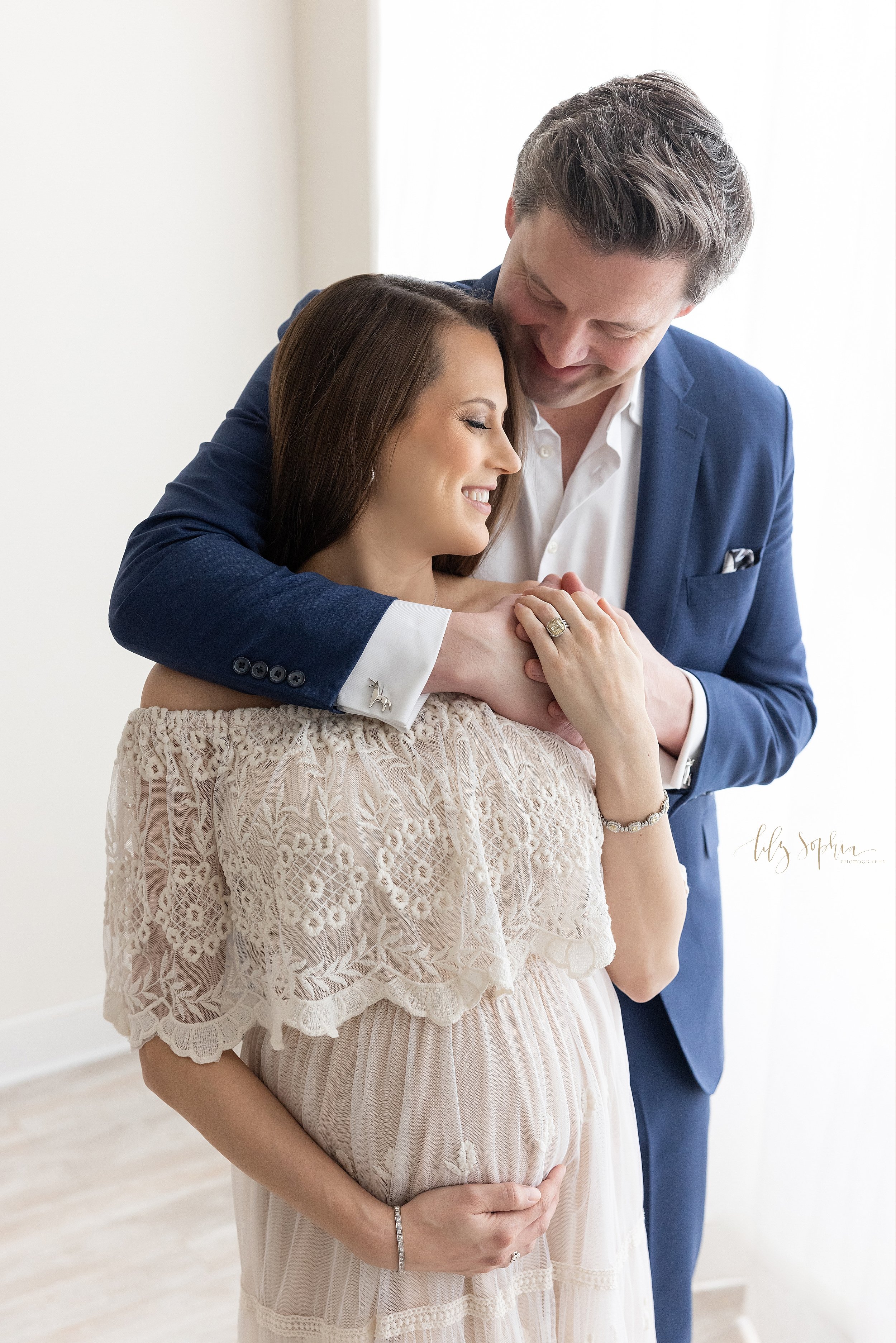  Maternity photo session with a husband wrapping his arms around his wife as she stands with her back to him and looks over her left should with her left hand holding on to her husband’s hands and her right hand framing the base of her belly as the c
