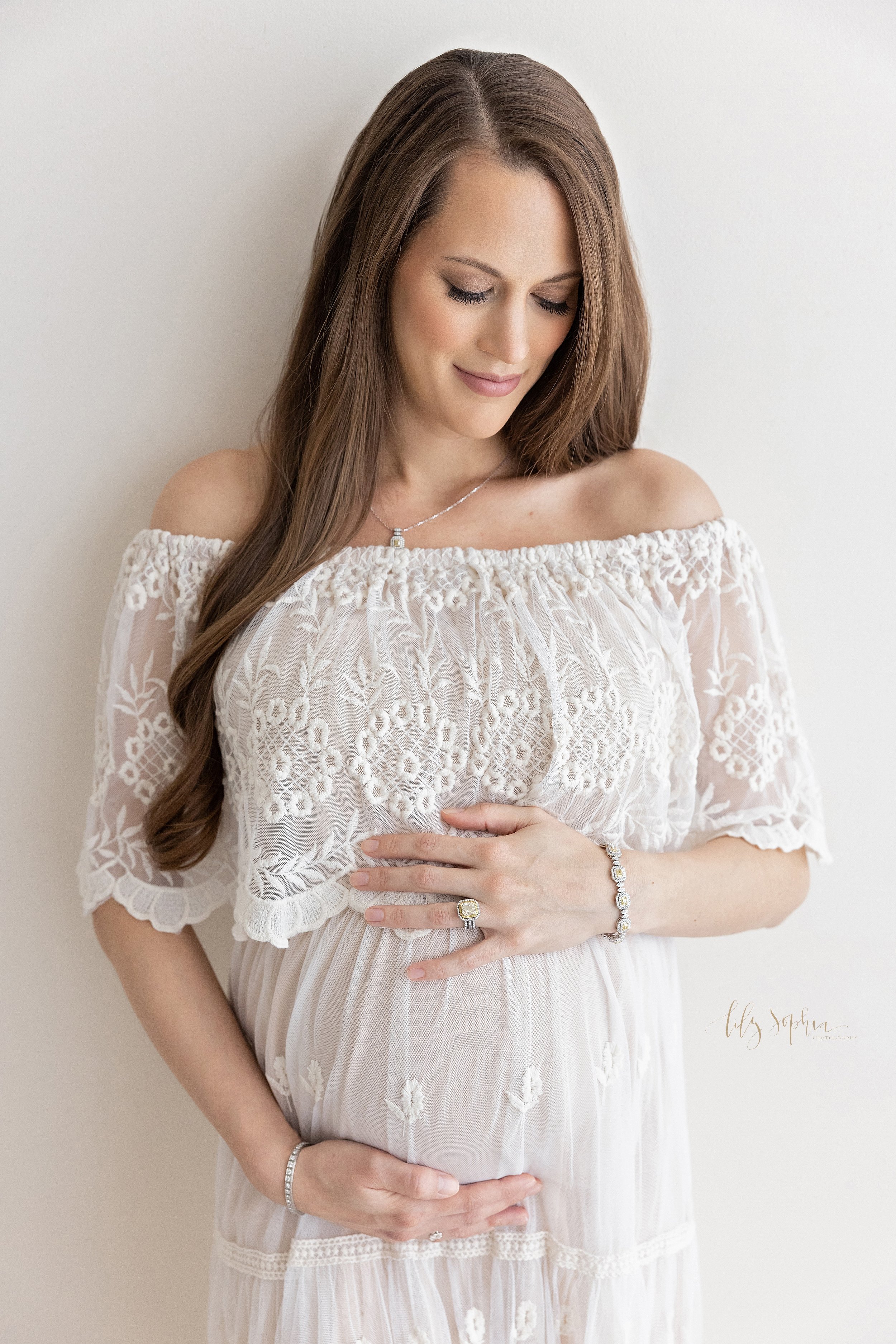  Maternity portrait of a contemplative pregnant mother wearing an off-the-shoulder full-length gown with an embroidered bodice as she stands against a wall framing her belly with her hands taken in a studio near Brookhaven in Atlanta that uses natura