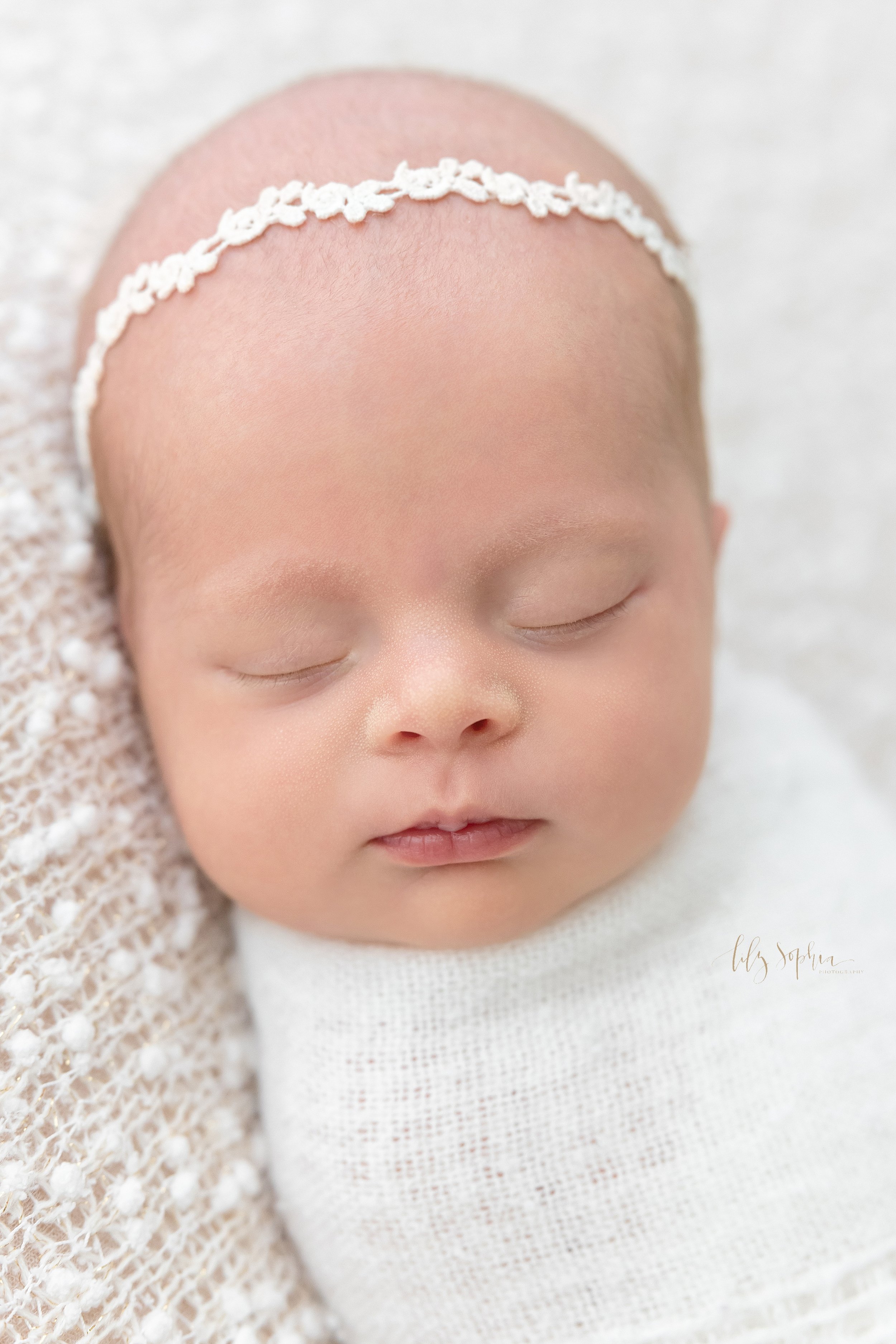  Close-up newborn portrait of a peacefully sleeping newborn baby girl wearing a delicate laurel headband in her hair and swaddled to her chin in a soft white blanket as she lies on her back taken in a natural light studio near Sandy Springs in Atlant