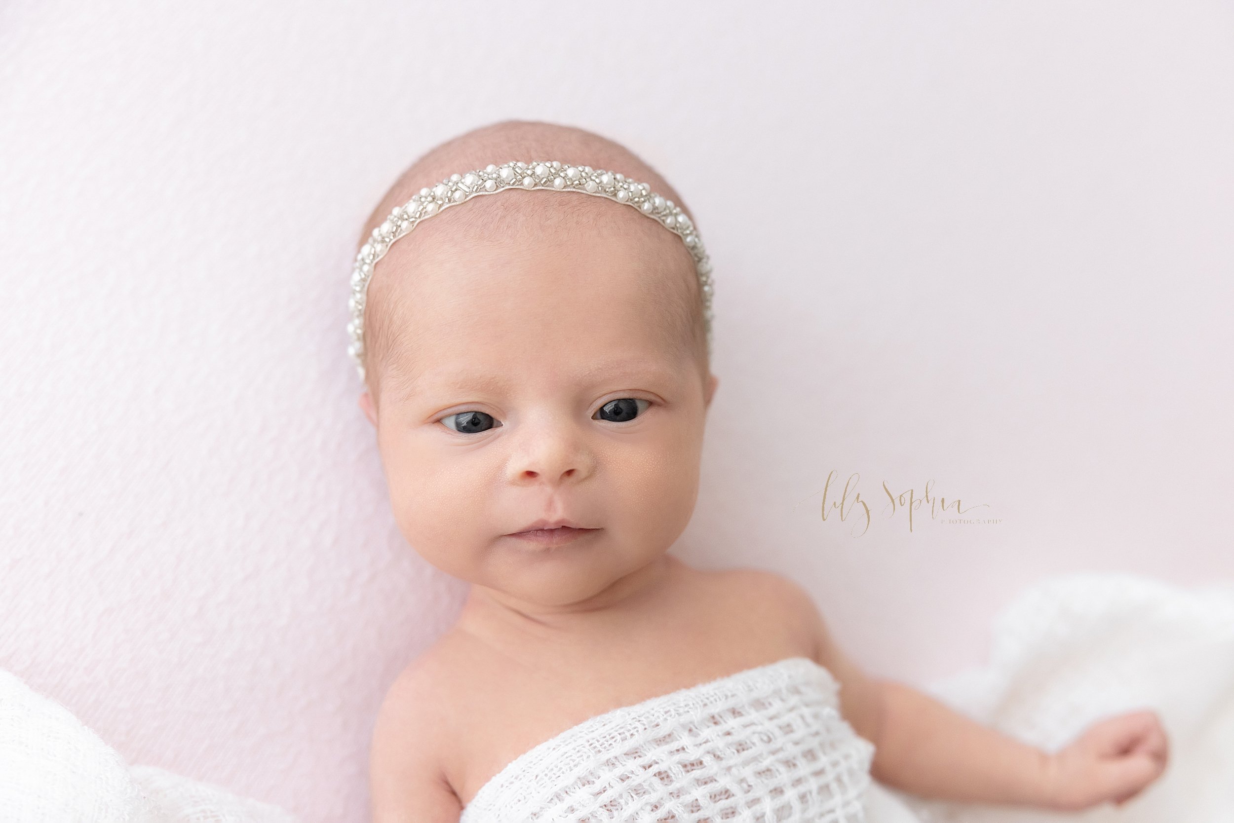  Newborn portrait of a wide awake newborn baby girl wrapped in a soft white knitted blanket and wearing a pearl headband on her head taken in a studio near Kirkwood in Atlanta that uses natural light. 