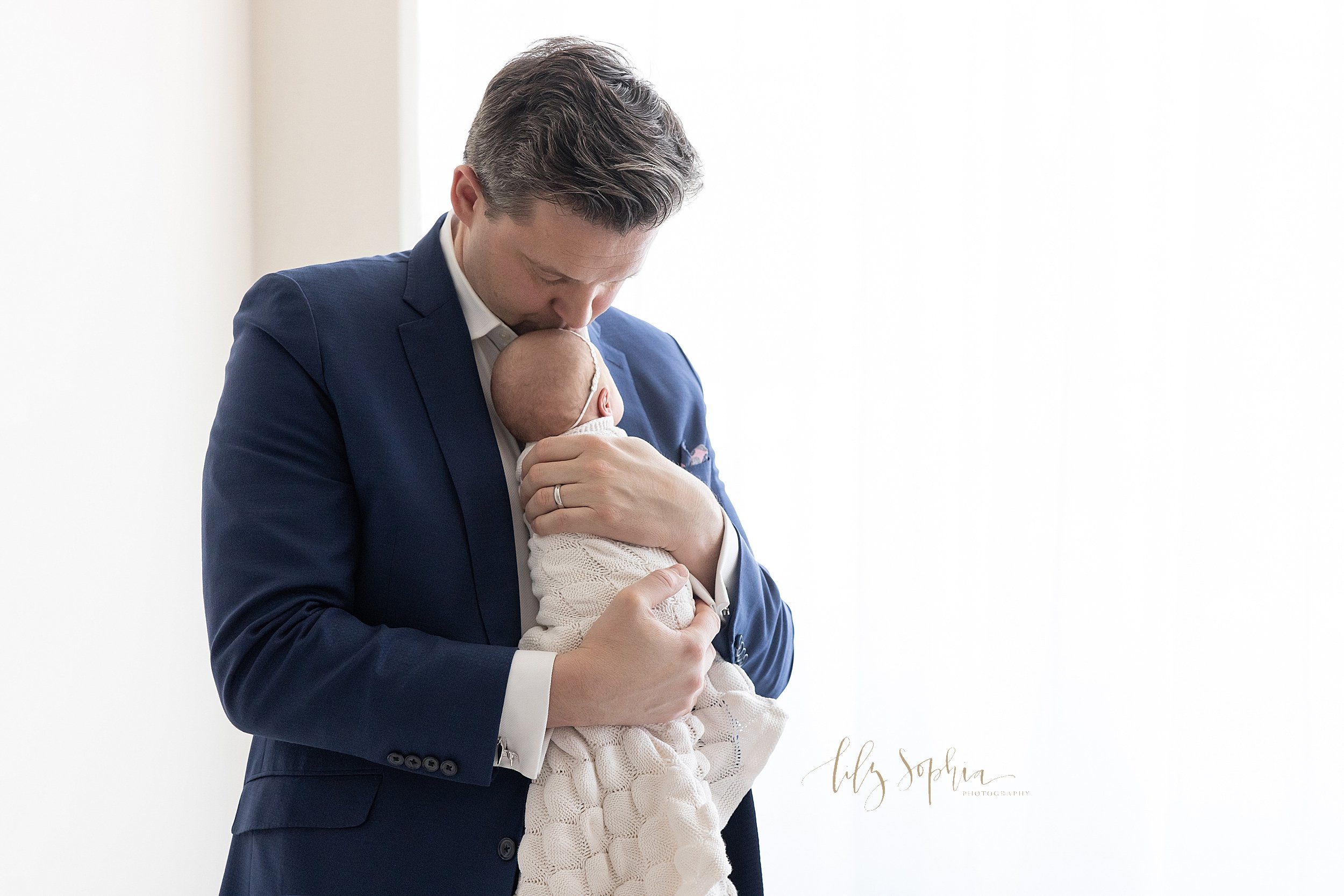  Newborn portrait of a father standing in front of a window streaming natural light in a studio holding his newborn daughter against his chest and kissing the crown of her head taken near Oakhurst in Atlanta. 