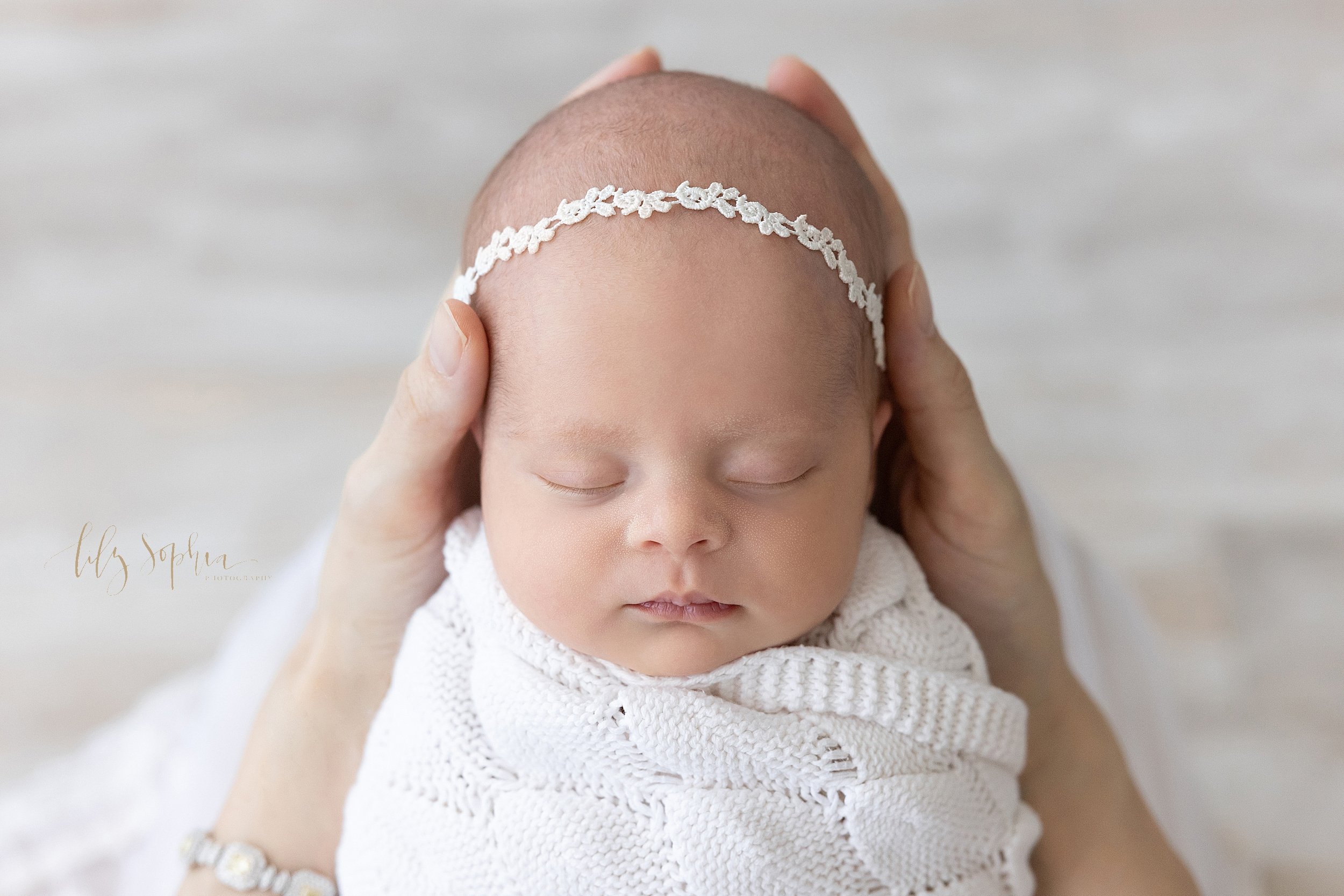  Newborn portrait of a newborn baby girl wearing a delicate laurel headband on her head as her head is held in her mother’s hand taken in Ponce City Market in Atlanta in a natural light studio. 