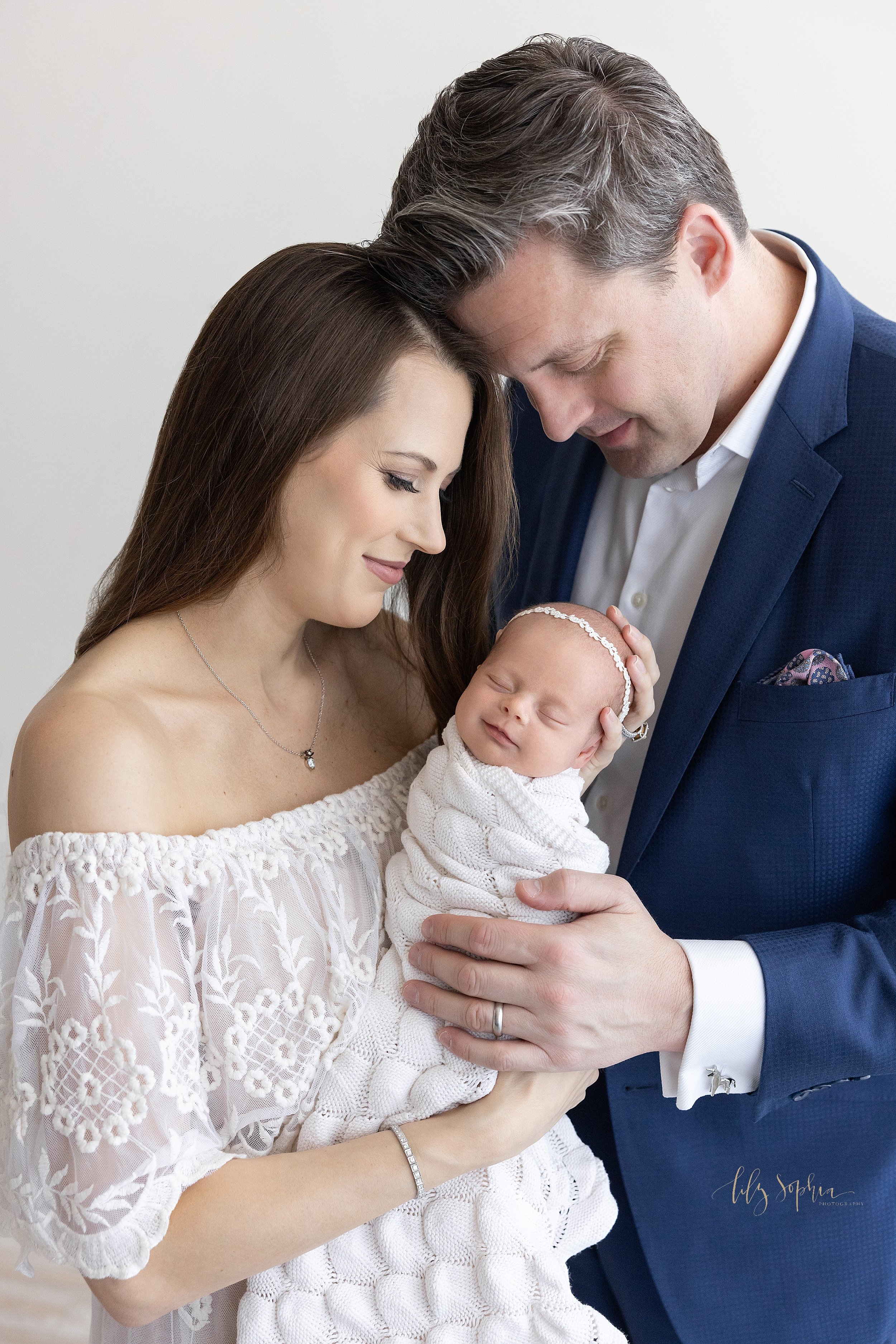  Newborn photo shoot with mom holding her peacefully sleeping and smiling newborn daughter who is swaddled in a soft white knit blanket to her chin and wearing a delicate laurel headband in her hair as dad stands next to mom’s left shoulder, places h