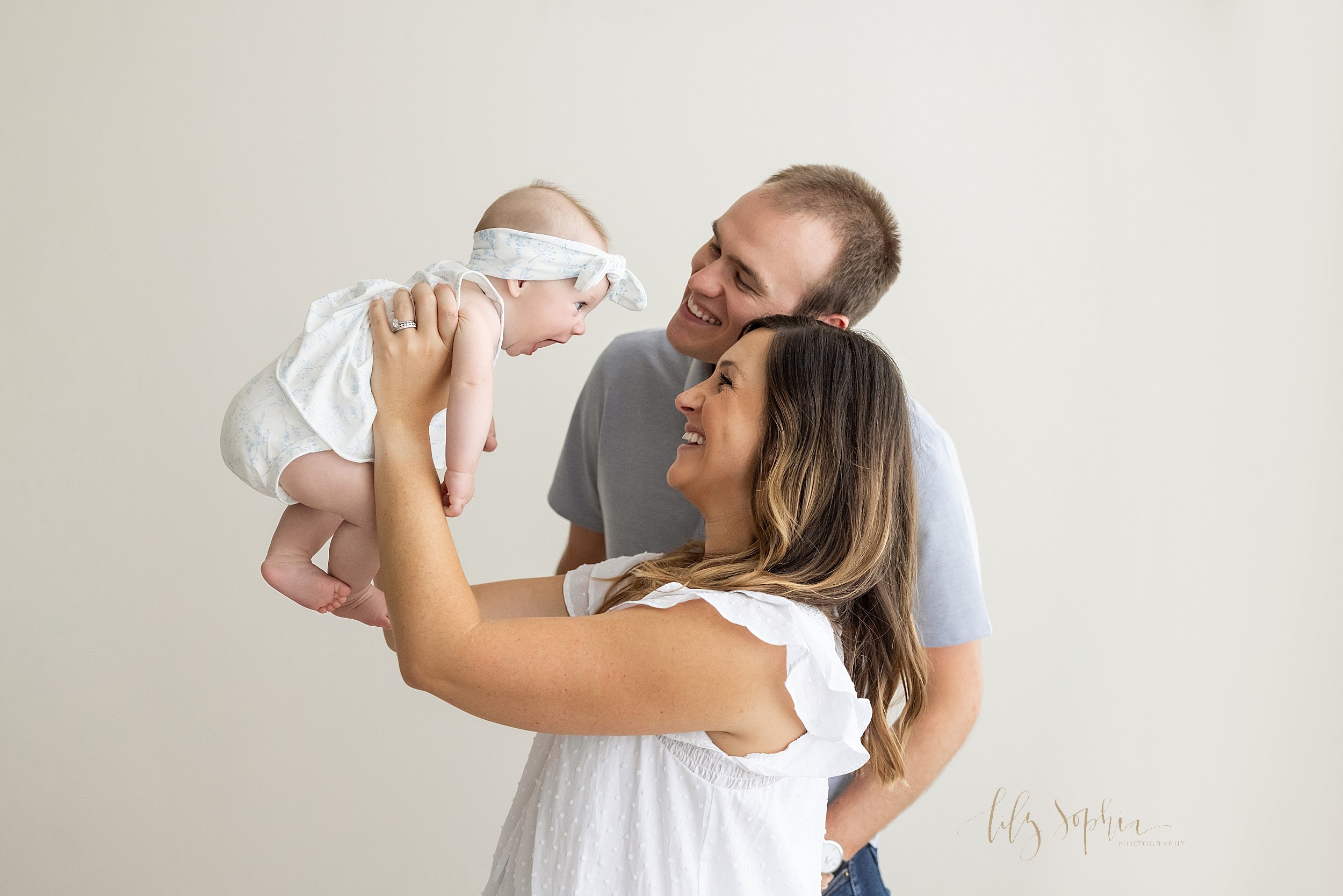  Family photo of a mother holding her baby girl above her head with dad standing on the right side of mom looking on as they interact with their daughter taken in a natural light studio near Brookhaven in Atlanta, Georgia. 