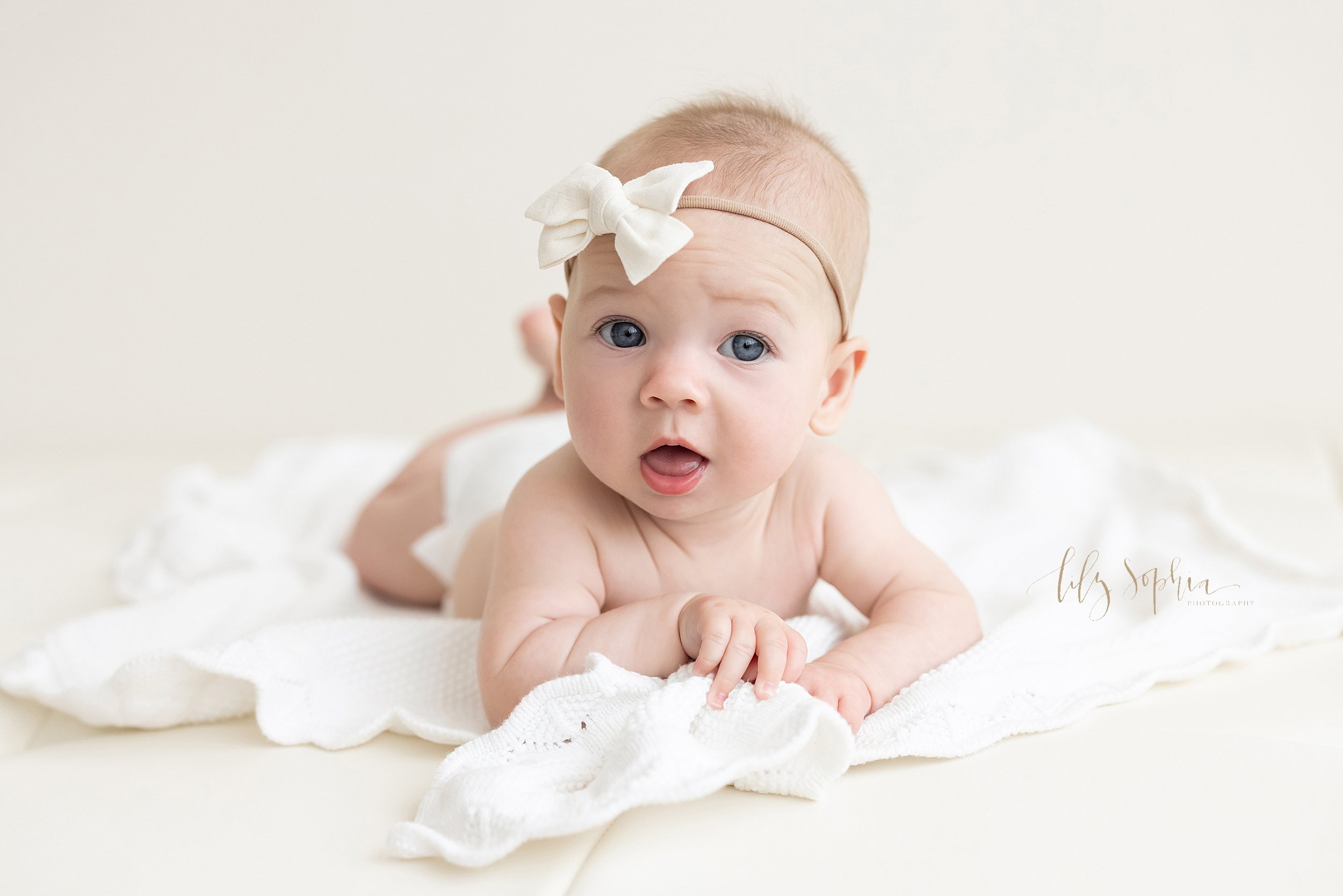  Baby photo session of a baby girl wearing a bow headband on her head as she lies on her stomach on a blanket taken in a studio near Ansley Park in Atlanta that uses natural light. 