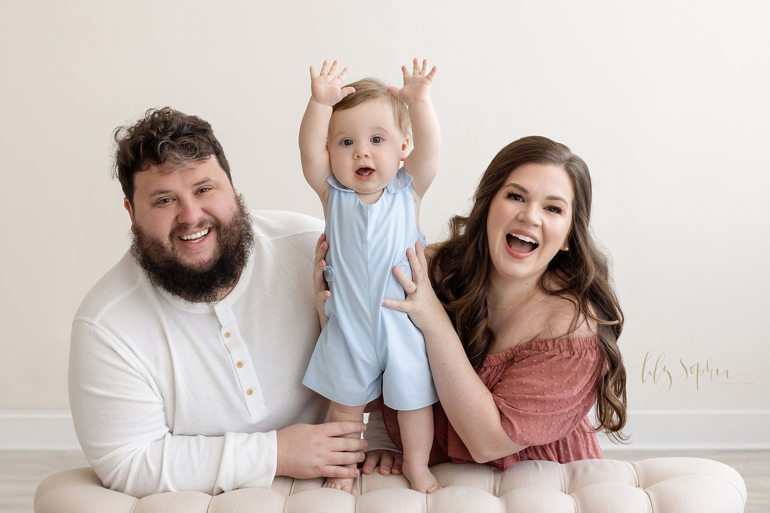  Family first birthday photo of a father and a mother kneeling in a studio behind a tufted bench with their one-year old son standing on the bench between them as he raises his hands to show how big he is taken near Sandy Springs in Atlanta using nat