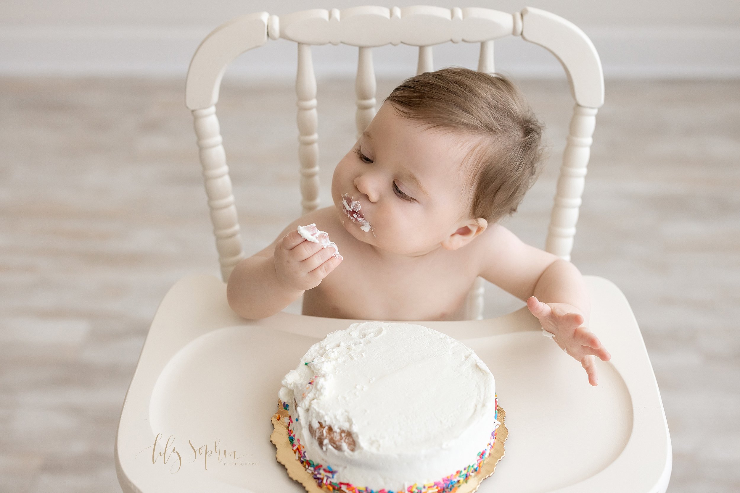  First birthday smash cake photo of a one-year old baby boy sitting in a wooden antique high chair in a natural light studio as he examines the bite of his smash cake in his hand taken near Morningside in Atlanta, Georgia. 