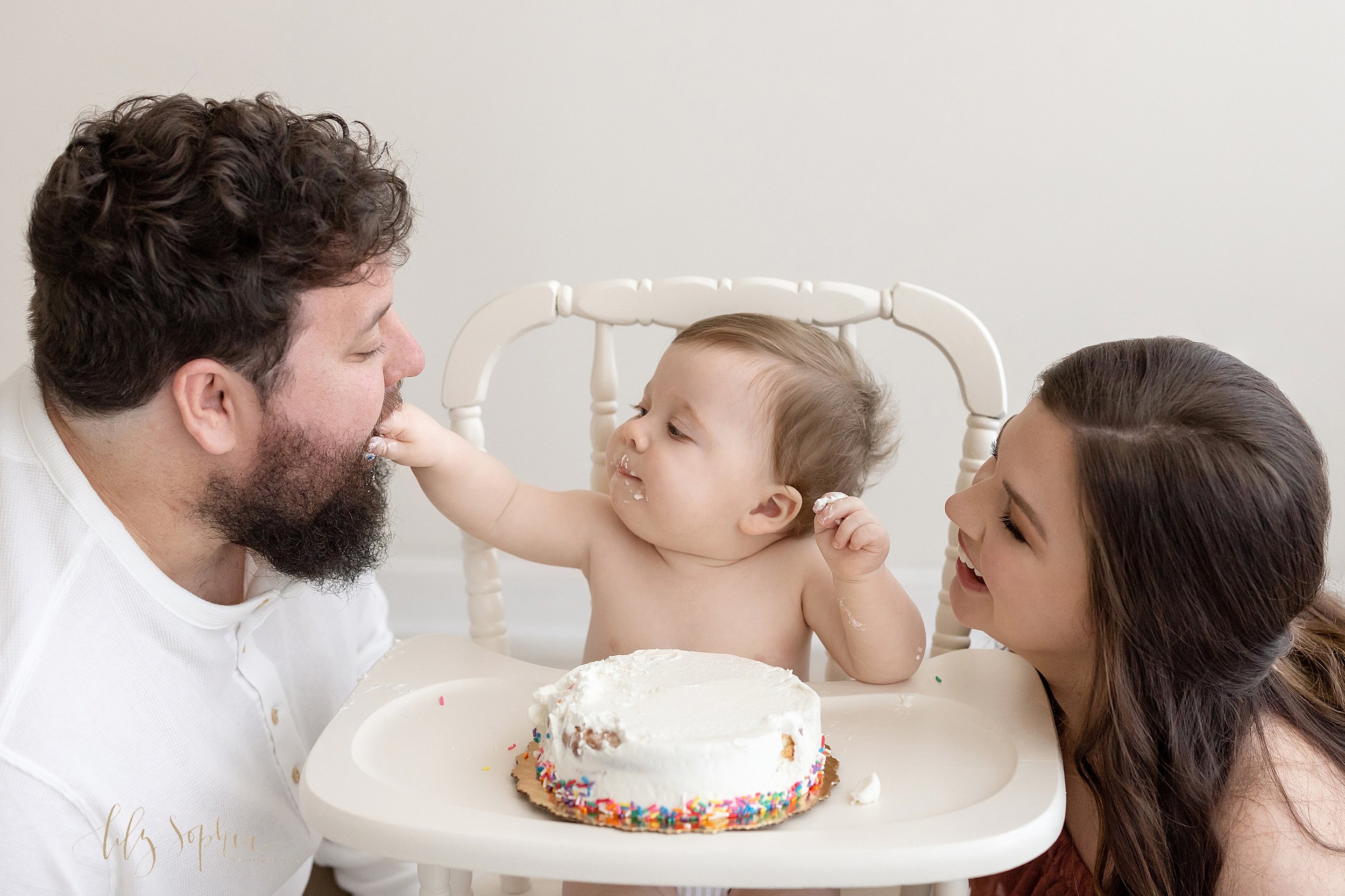  First birthday family photo shoot of a one-year old baby boy sitting in an antique high chair with his mother on his left side and his father on his right side as he feeds his dad a bite of his smash cake taken near Midtown using natural light in a 