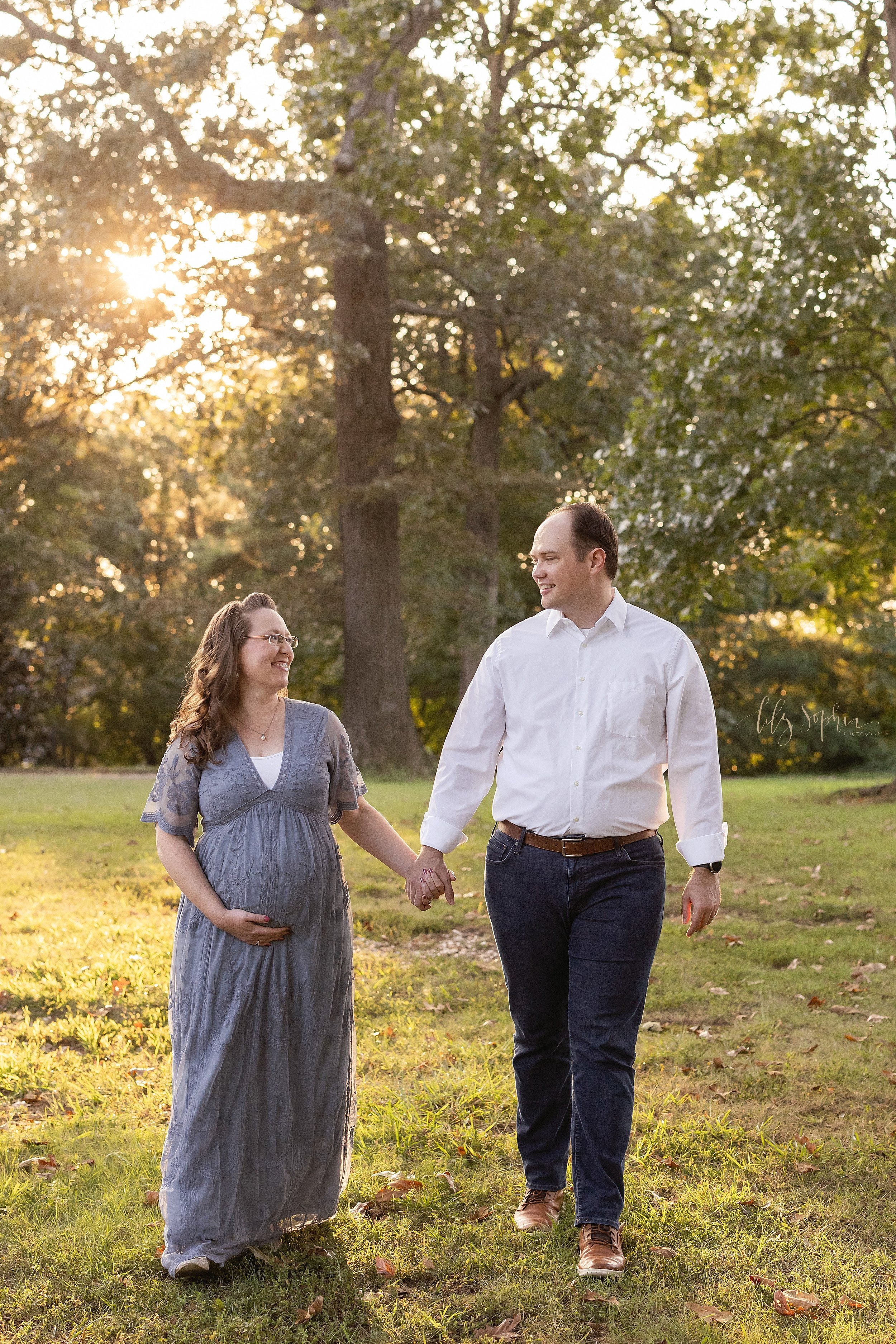  Maternity pictures with a pregnant mother holding the base of her belly with her right hand as she holds her husband’s hand with her left hand as the couple walks at sunset in an Atlanta, Georgia park. 