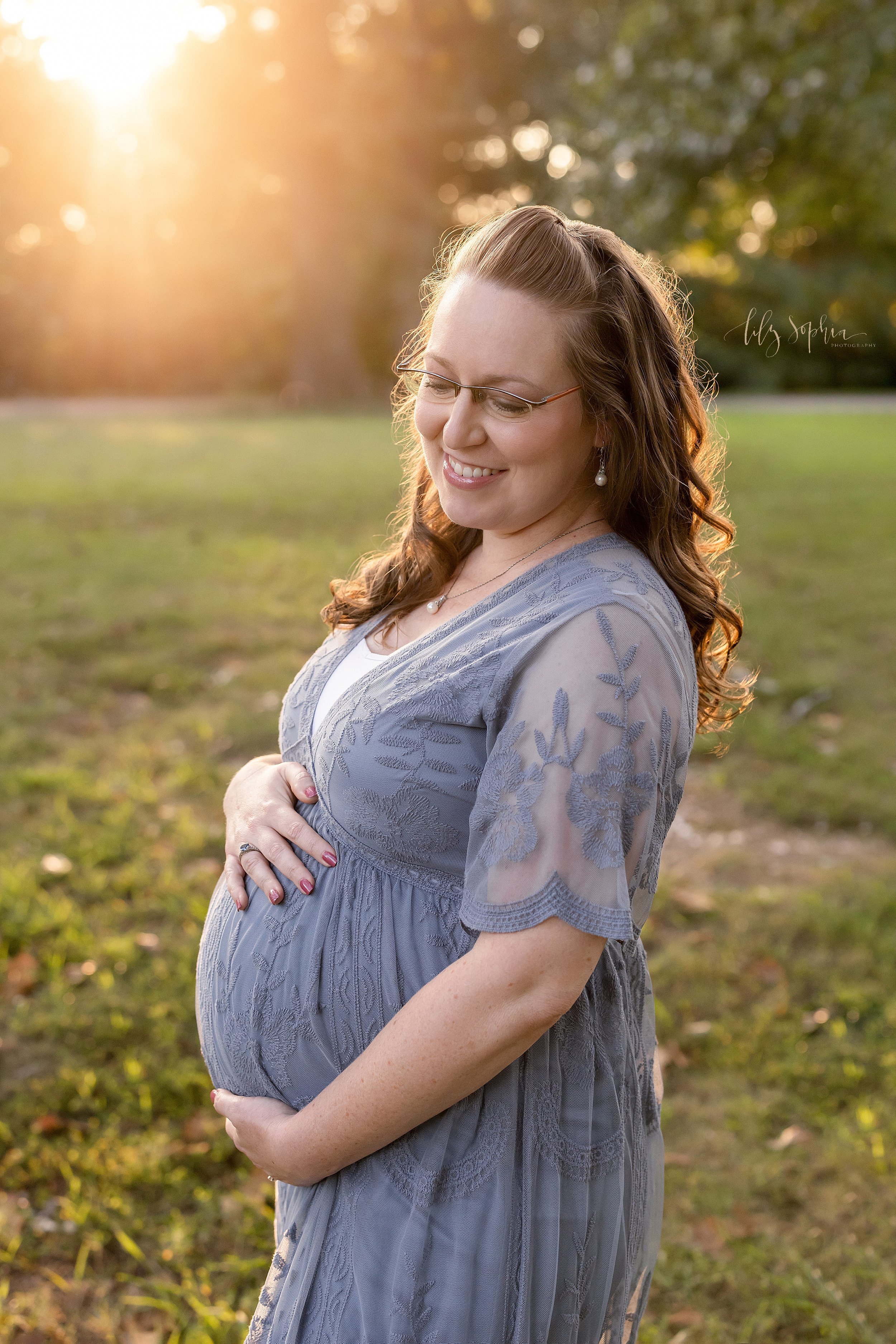  Maternity portrait of a pregnant mother as she stands in an park with her hands holding her baby in utero at sunset near Atlanta, Georgia. 