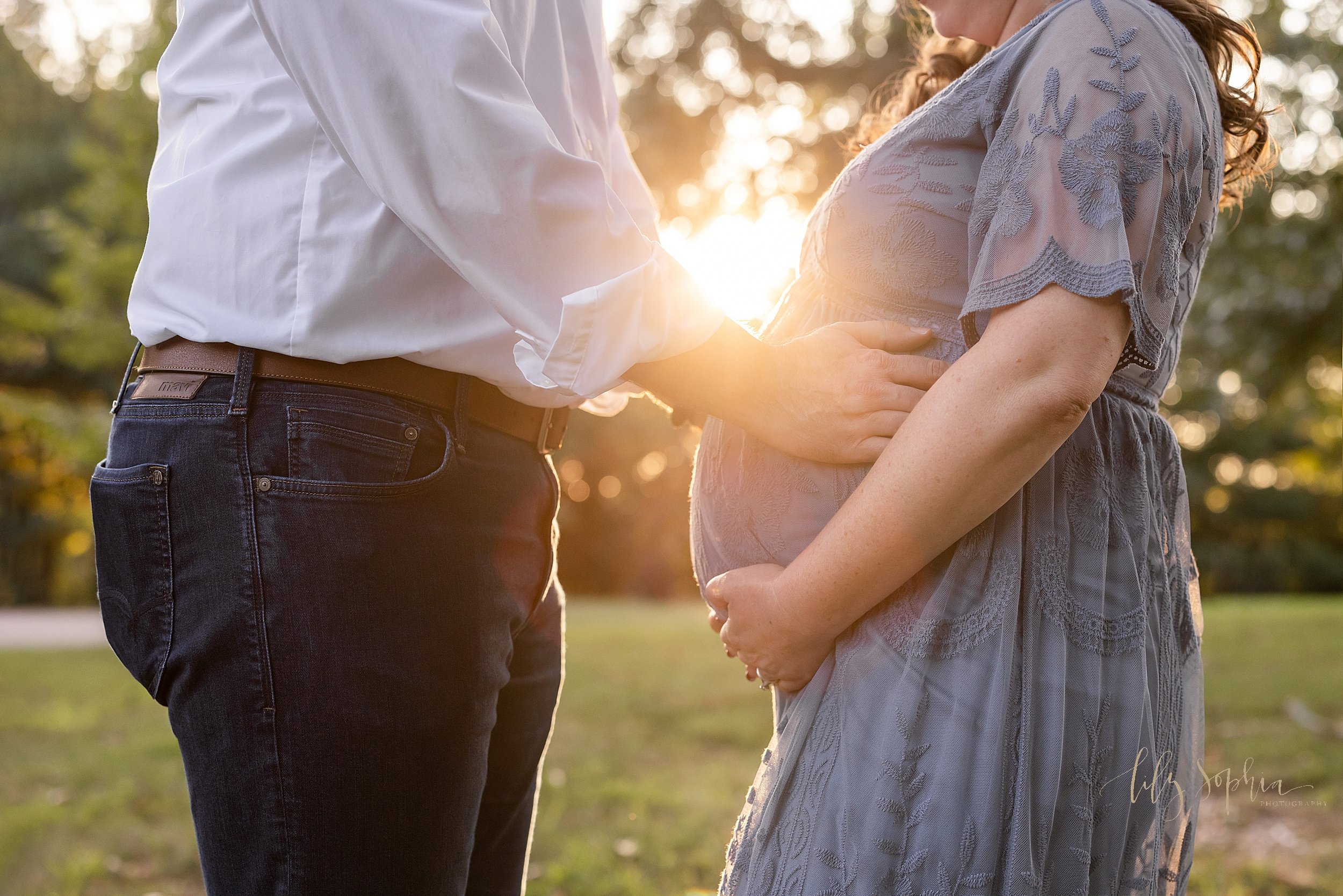  Close-up maternity photo of a husband facing his wife with his hands on his child in utero while the pregnant mother holds her child in utero with the sunset in the background as the couple stand in a park near Atlanta. 