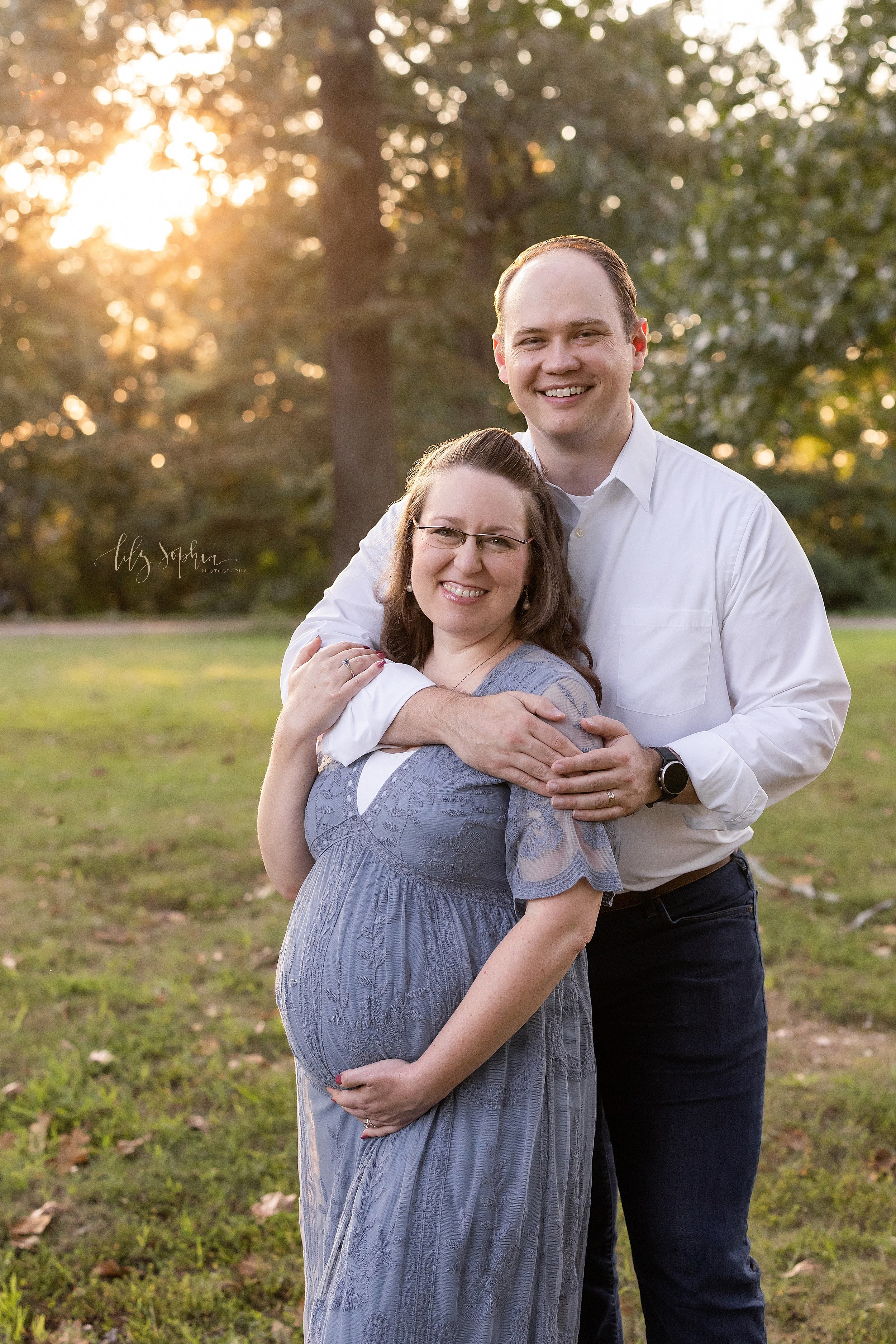  Maternity photo shoot in an Atlanta park with the husband standing behind his wife as he wraps his arms around her shoulders and she holds his arm with her right hand and frames her belly with her left hand as the sunset goes down in the background.