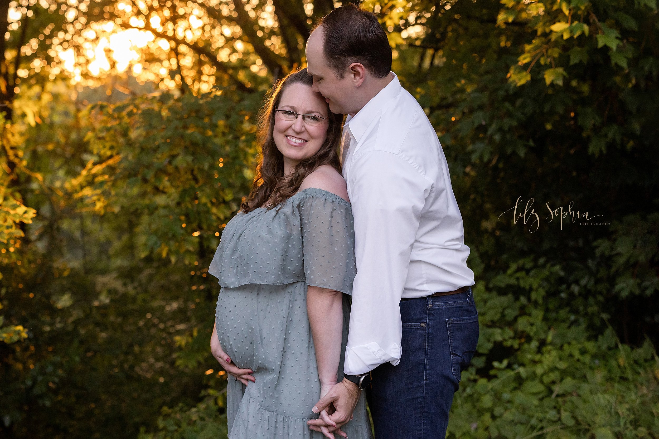  Maternity pictures of a husband standing behind his wife and holding her left hand in his as he kisses the top of his wife’s head as she turns her head over her left shoulder and holds the base of her belly with her right hand taken at sunset in an 