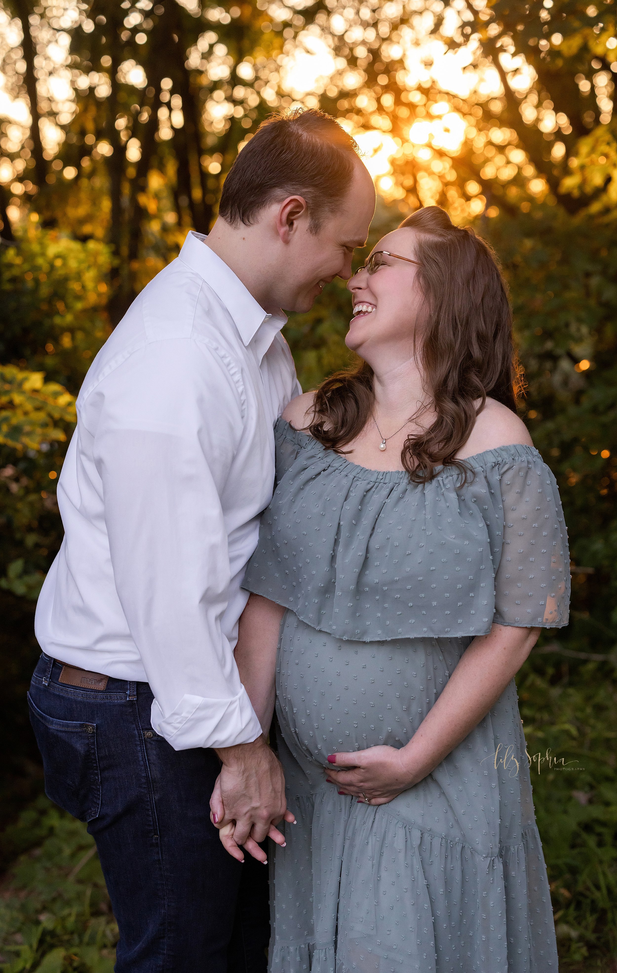  Maternity photo session in an Atlanta park at sunset with the husband holding his wife’s right hand with his left hand while standing to her right side and the wife holding the base of her belly and the two of them get ready to kiss. 