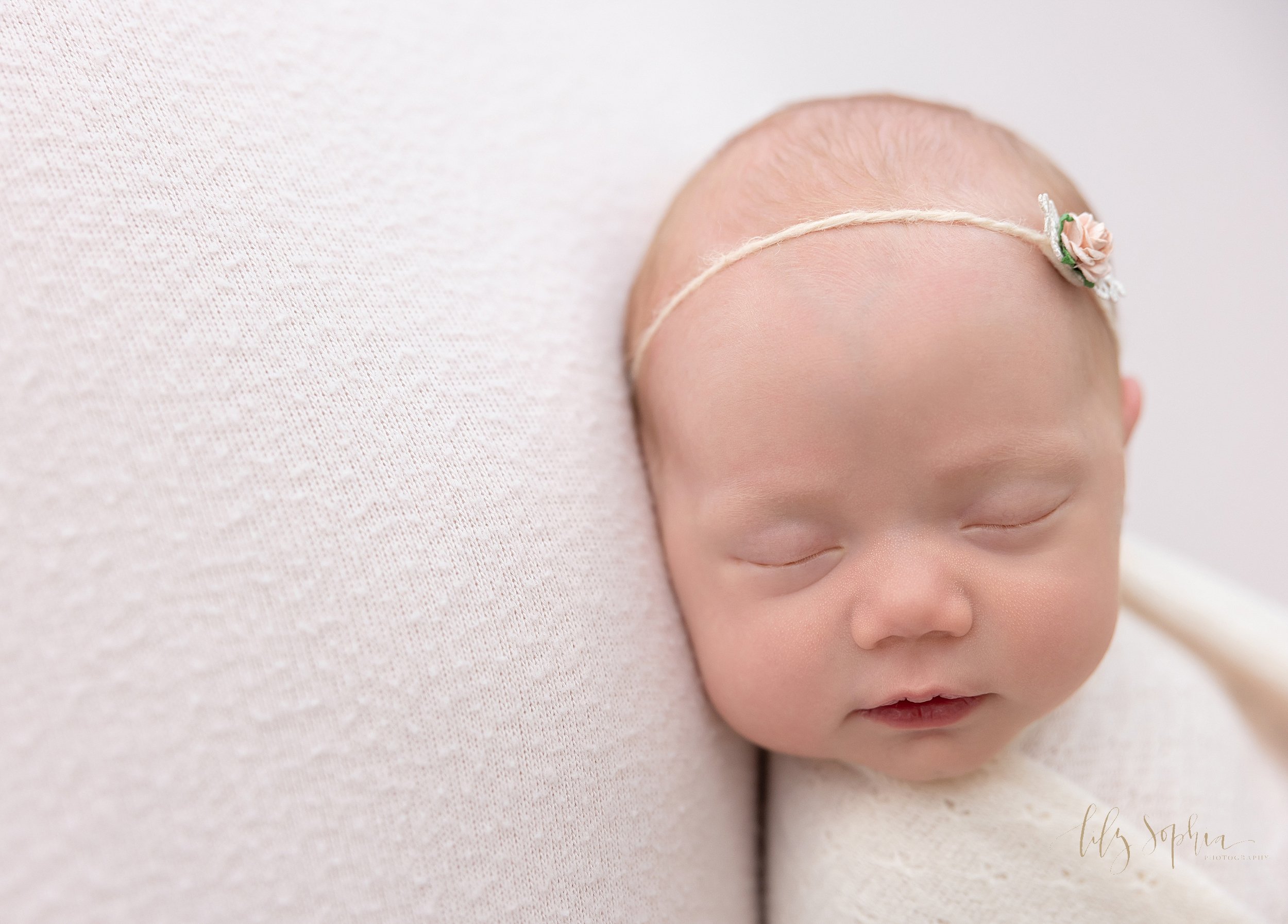  Newborn photo shoot of a beautiful newborn baby girl as she lies on her back wearing a headband adorning a single rose as she is swaddled to her chin with a soft white blanket taken in a natural light studio near Old Fourth Ward in Atlanta, Georgia.