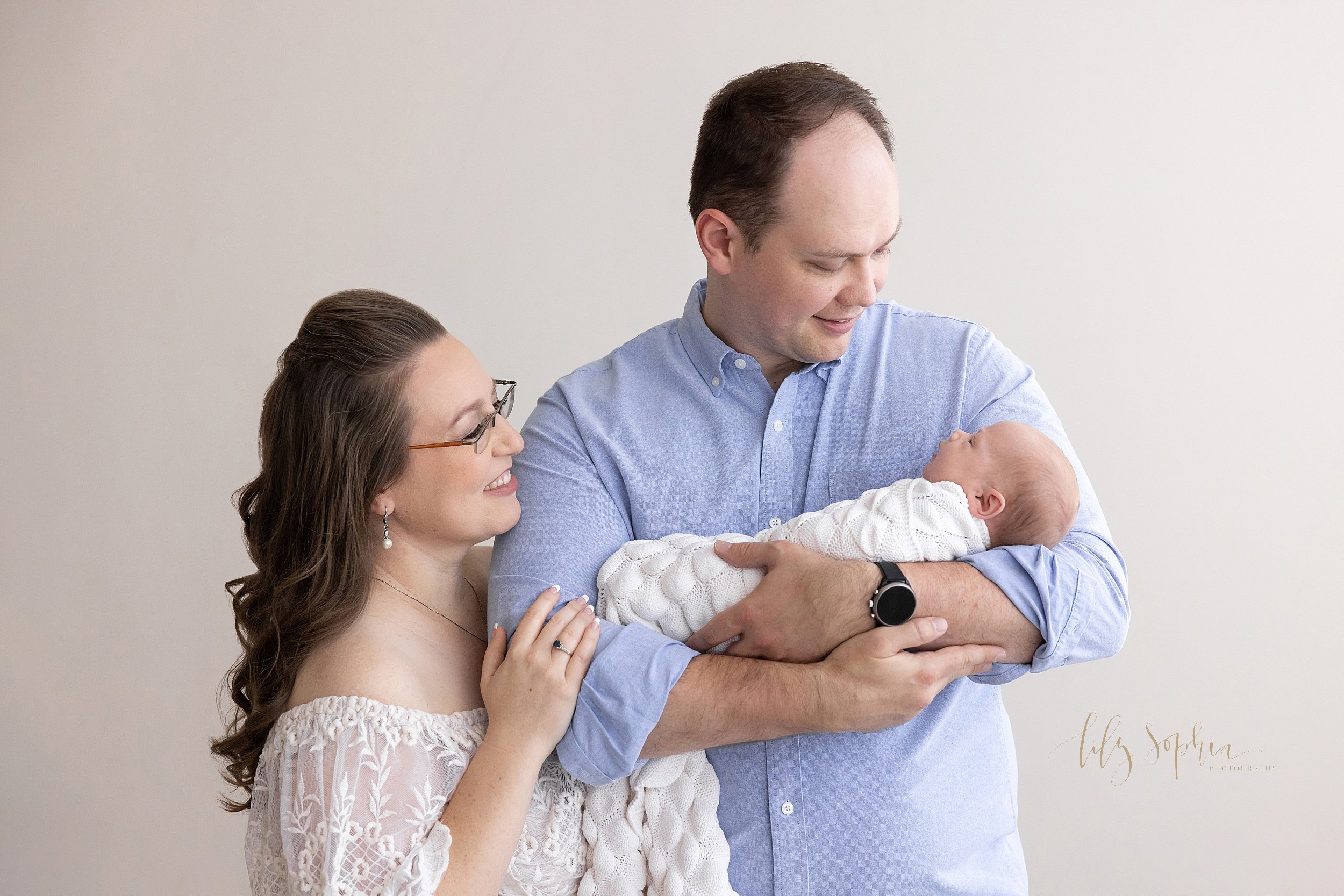  Newborn family portrait of a father cradling his peacefully sleeping newborn daughter in his arms while the baby’s mother stands on the right side of the father proudly looking on as she holds her husband’s right arm taken in a natural light studio 