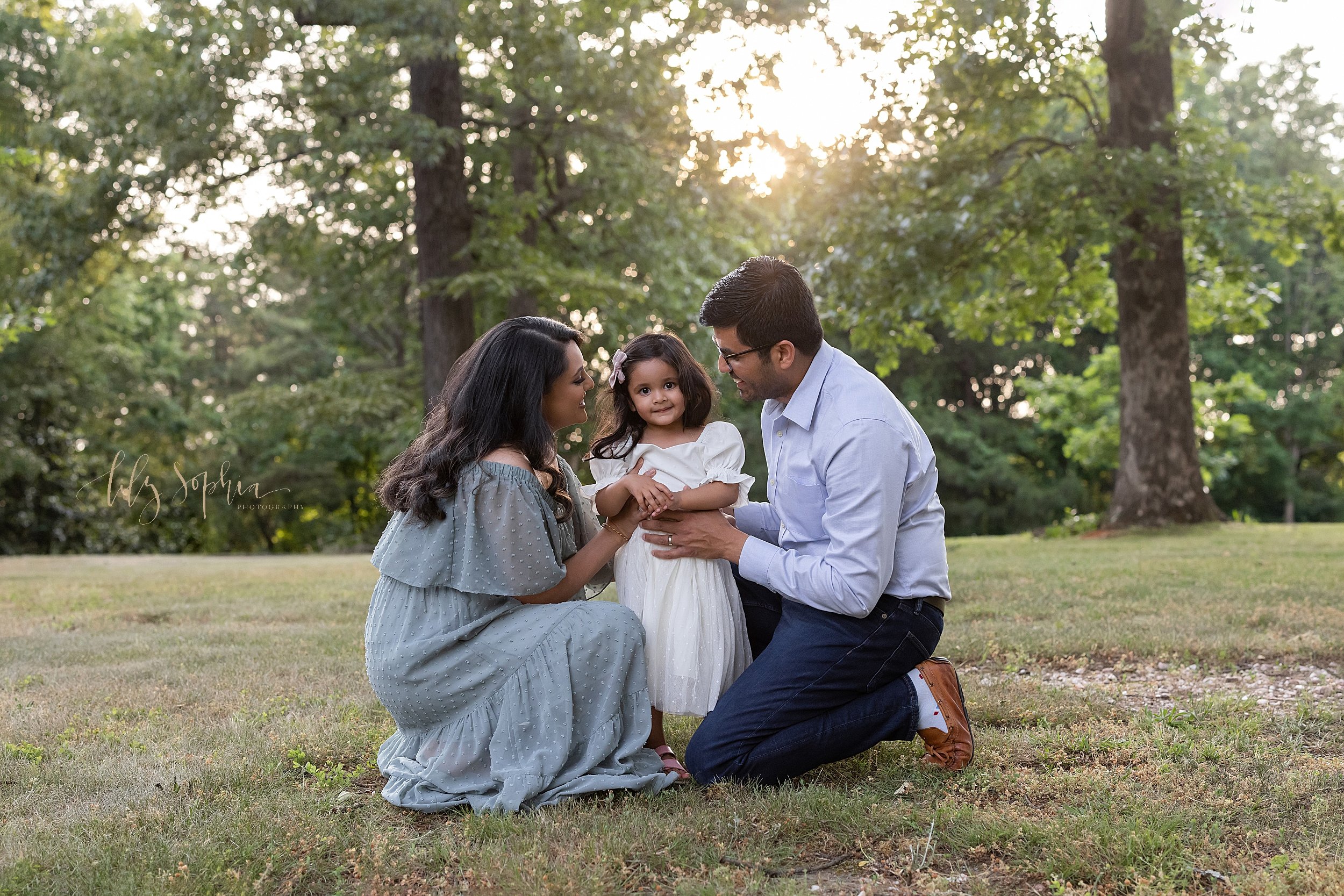  Family photo session in an Atlanta park of an Indian mother and father squatting with their daughter between them while the sun sets behind them. 