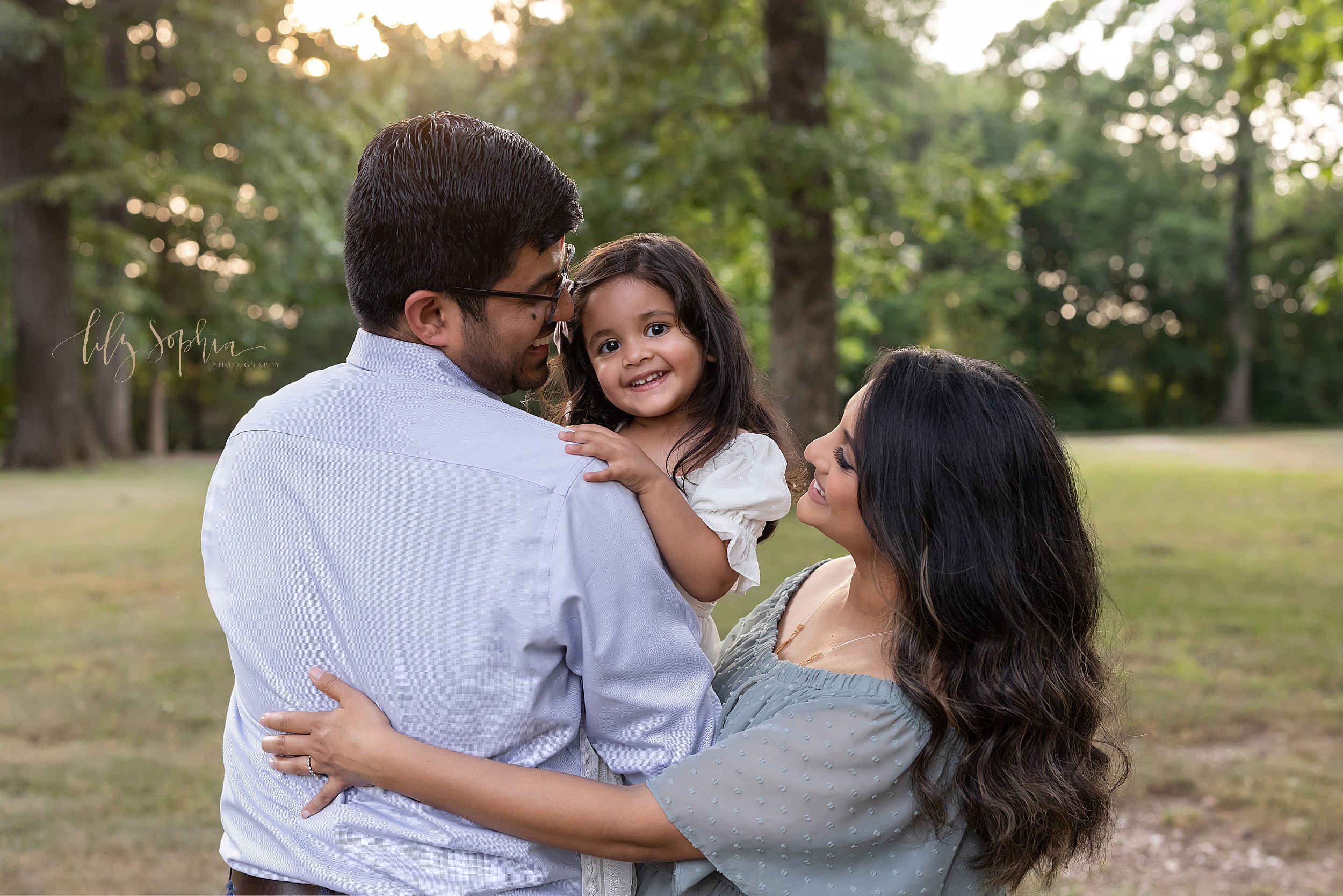  Family portrait of a father holding his young daughter in his arms as she peeks over his right shoulder and his wife stands to his right side with her arm on his back taken in an Atlanta park at sunset. 