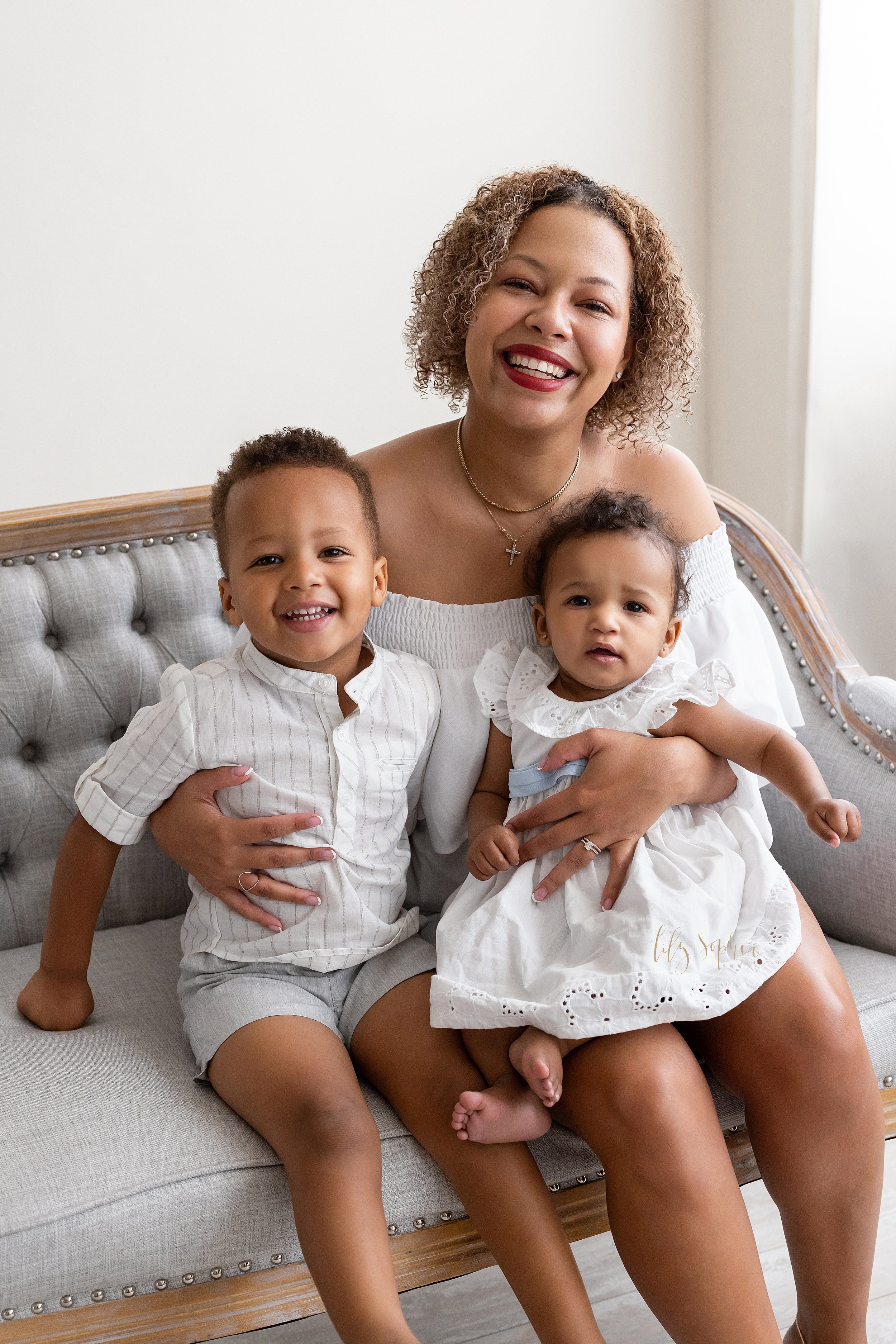  Family portrait of an African-American mother sitting with her baby girl on her lap and her young son next to her as they sit on a tufted sofa in front of a window streaming natural light in a studio near Oakhurst in Atlanta, Georgia. 