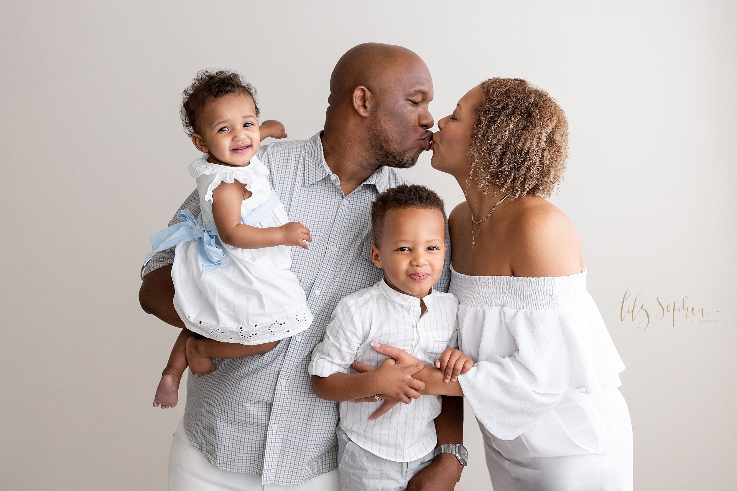  Family portrait of an African-American family with dad holding their baby girl in his right arm as he turns to kiss his wife who is on his left side as she holds onto their son who is standing between them taken in a natural light studio near Midtow