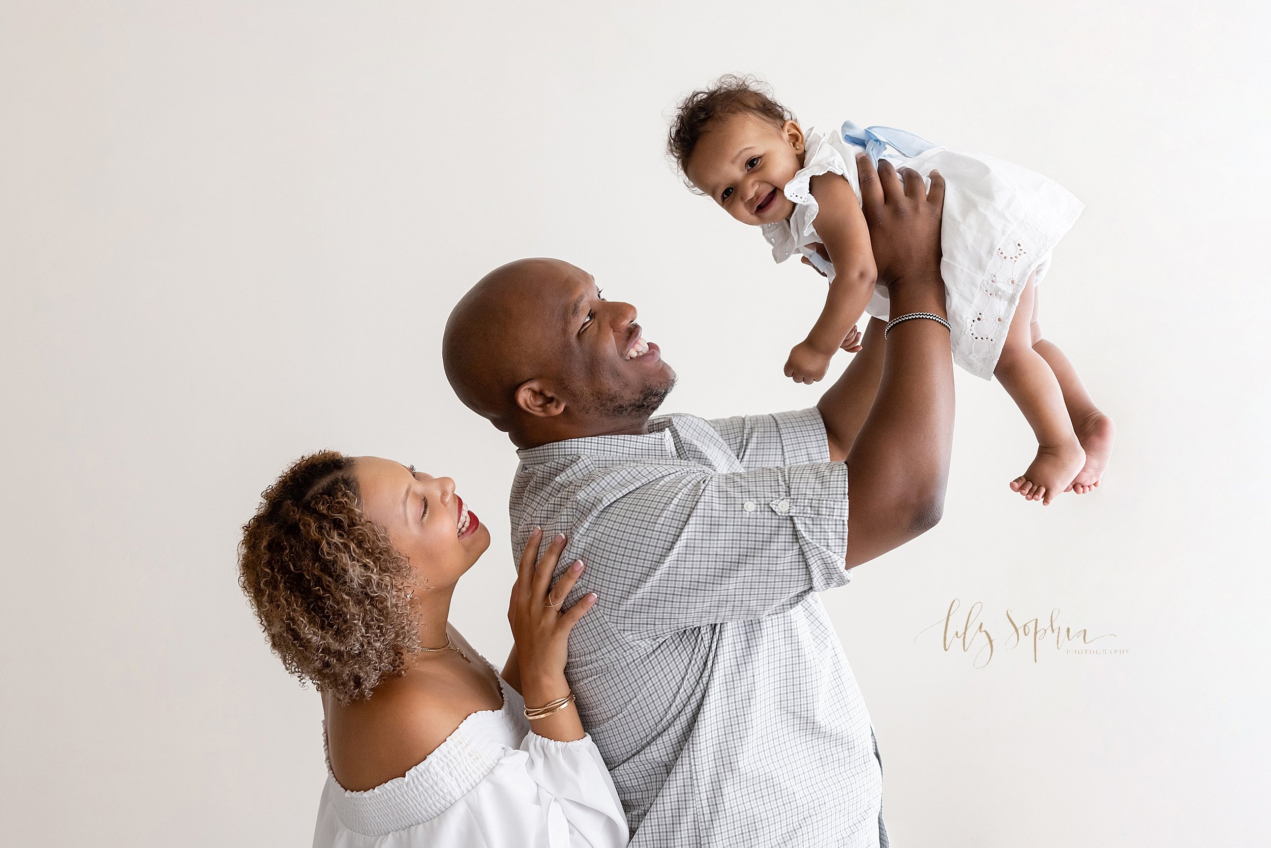  Baby photo of a baby girl being held in her fathers arms as he raises her above his head and his wife stands behind him with her hand on his back taken in front of a window streaming natural light in a studio near Decatur in Atlanta, Georgia. 