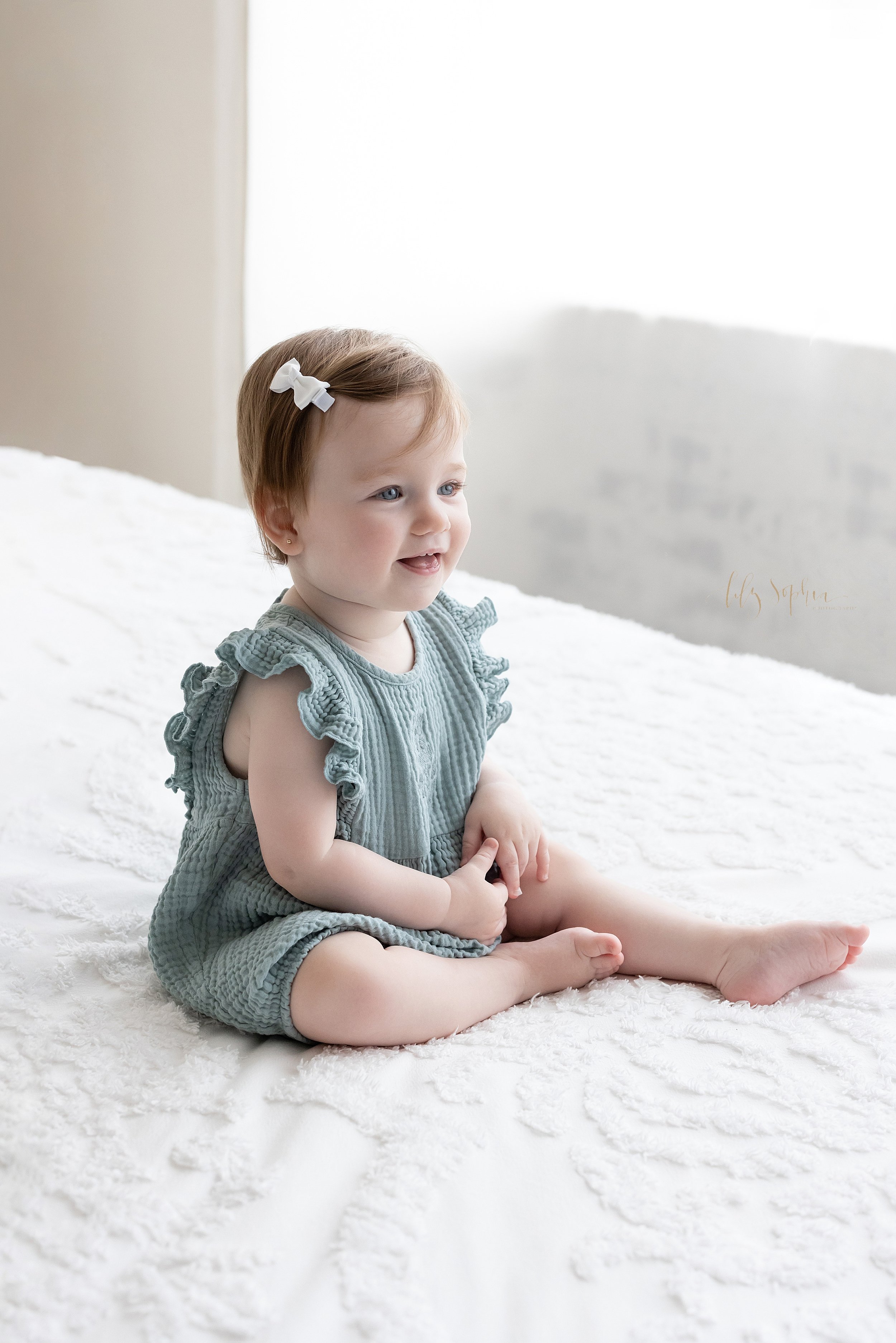  First birthday portrait of a one year old little girl as she sits on top of a bed in front of a window streaming natural light in a studio near Buckhead in Atlanta. 