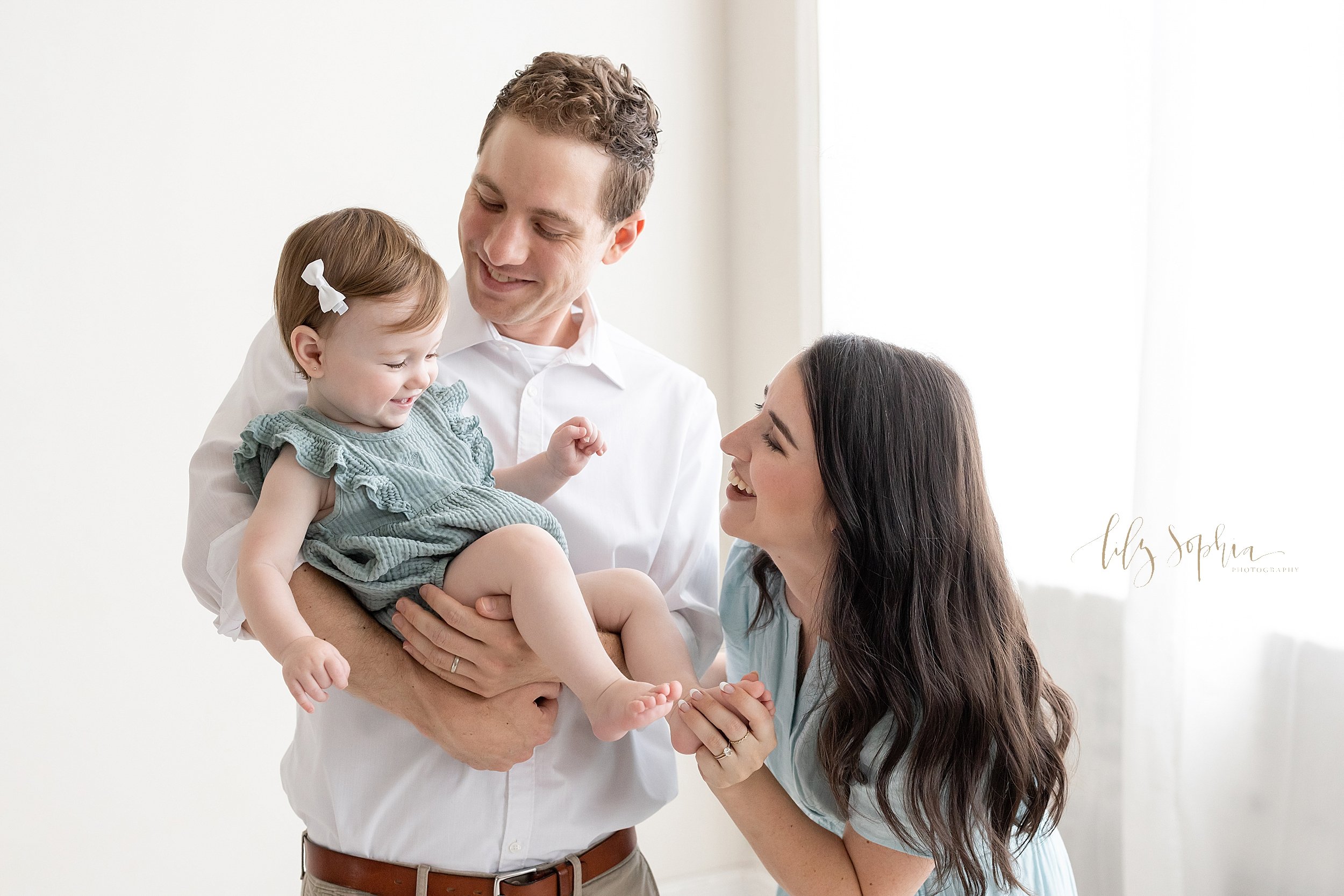  First birthday family photo session of a father standing in a natural light studio holding his one year old daughter in his arms as mom stands next to dad and tickles her daughter’s foot taken near Oakhurst in Atlanta. 