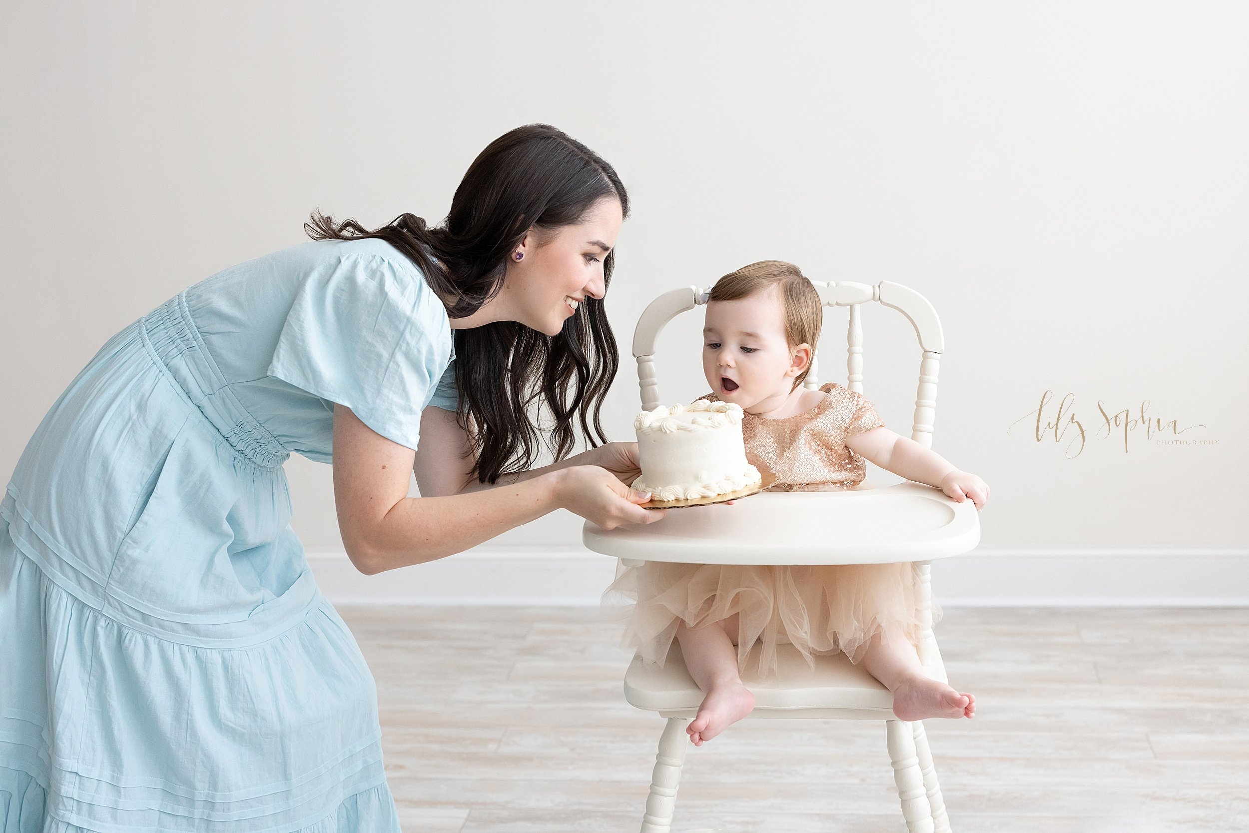  First birthday smash cake photo shoot of a one year old little girl sitting in an antique high chair as her mom brings her smash cake to place on the tray taken in natural light near Ansley Park in a photography studio. 