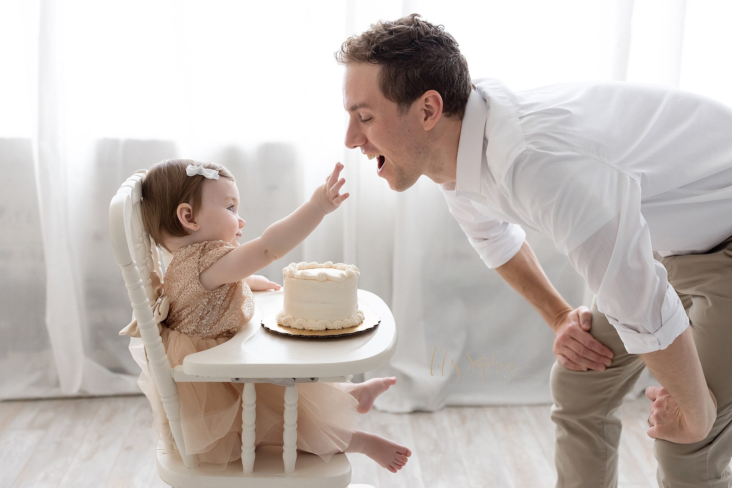  First birthday smash cake photo shoot of a one year old little girl giving a bite of her cake to her father as she sits in a high chair in front of a window streaming natural light near Kirkwood in Atlanta in a photography studio. 