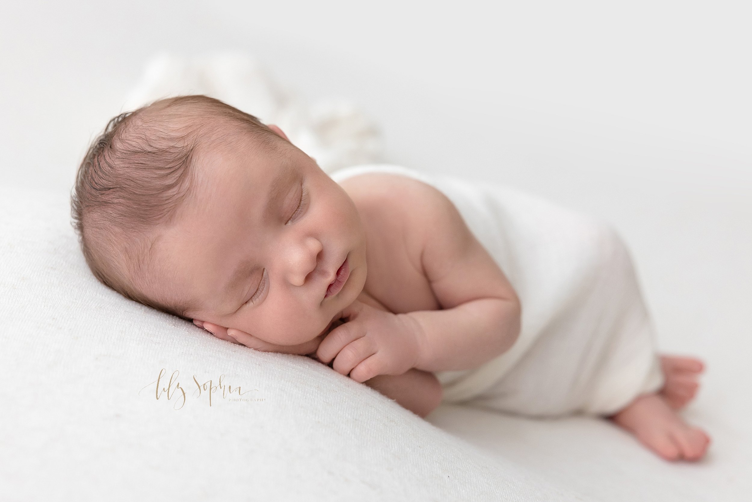  Newborn portrait of an infant boy as he sleeps on his side with his right hand under his cheek and his tiny feet sticking out of a soft white blanket taken near Sandy Springs in Atlanta in natural light in a photography studio. 