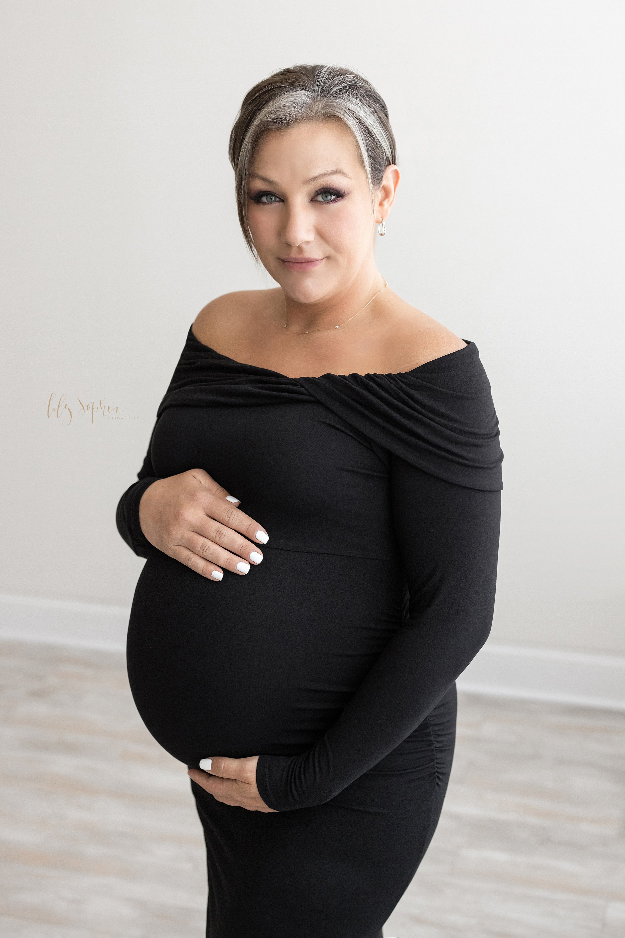  Maternity portrait of a pregnant mother wearing a black jersey knit long-sleeved gown as she stands and frames her belly with her hands taken in Ponce City Market in the Lily Sophia Photography Studio.. 