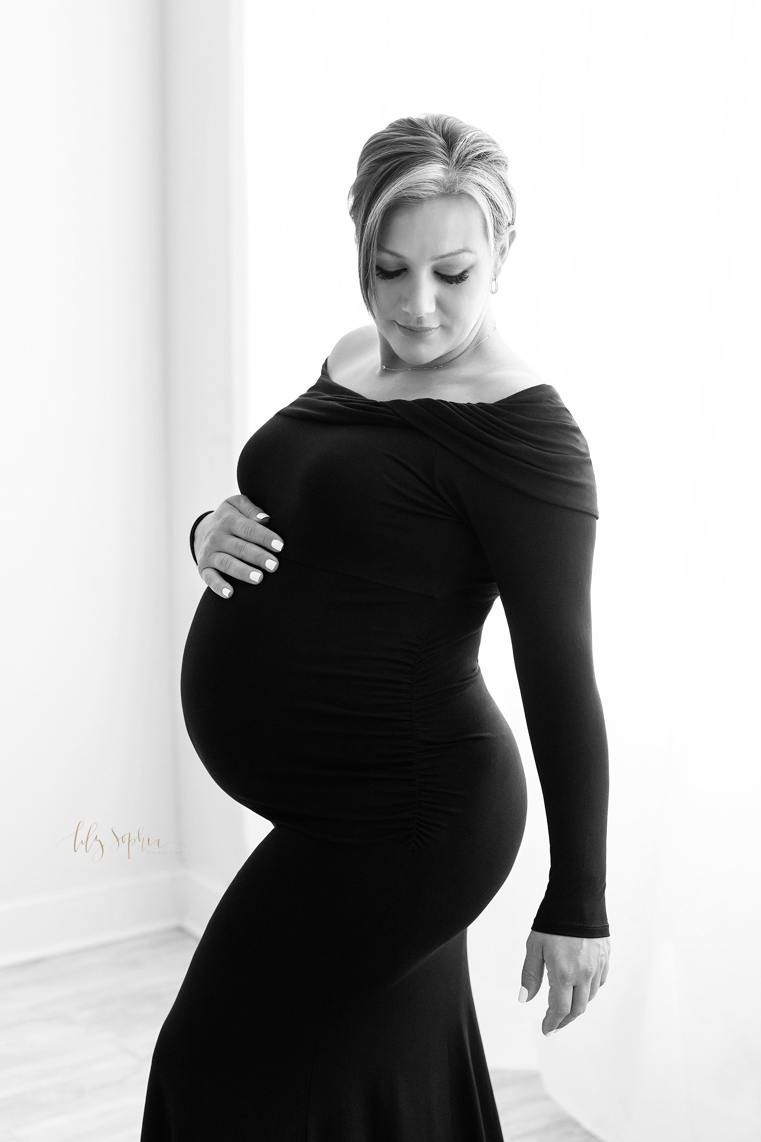  Maternity photo session of a pregnant mother wearing a jersey knit long sleep black gown as she stands in front of a natural light window with her right hand on the top of her belly taken near Midtown in Atlanta, Georgia in a photography studio. 