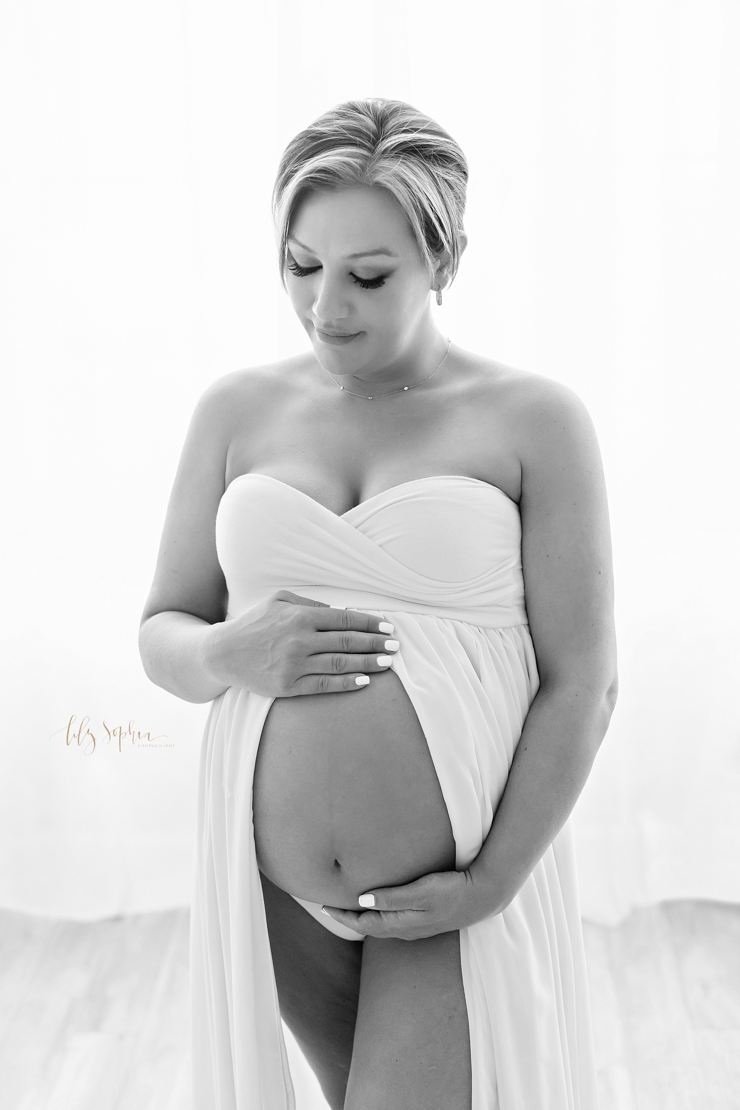  Maternity photo shoot of a pregnant mother wearing a strapless split front knit gown as she stands with her hands framing her bare belly taken in front of a window streaming natural light in a studio near Morningside in Atlanta. 