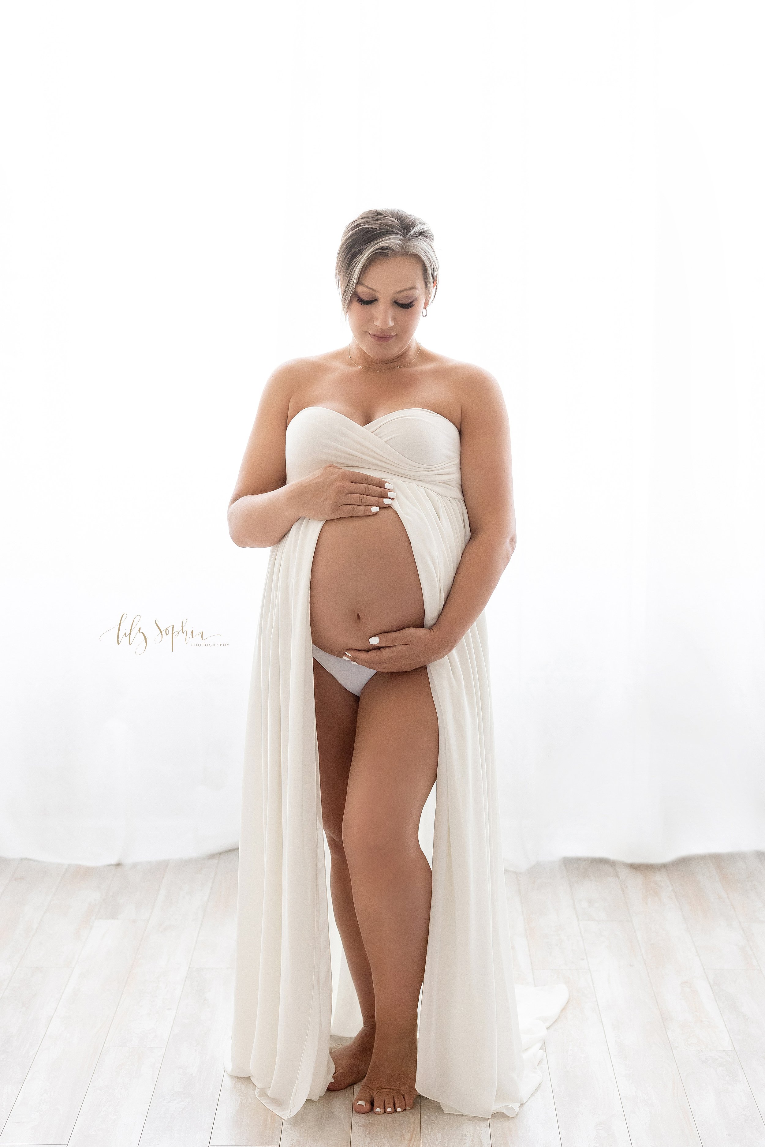  Maternity photo  shoot of a pregnant mother wearing a split front knit gown with her belly frame by her hands as she looks down and contemplates the birth of her child taken in front of a window streaming natural light in a photography studio near K