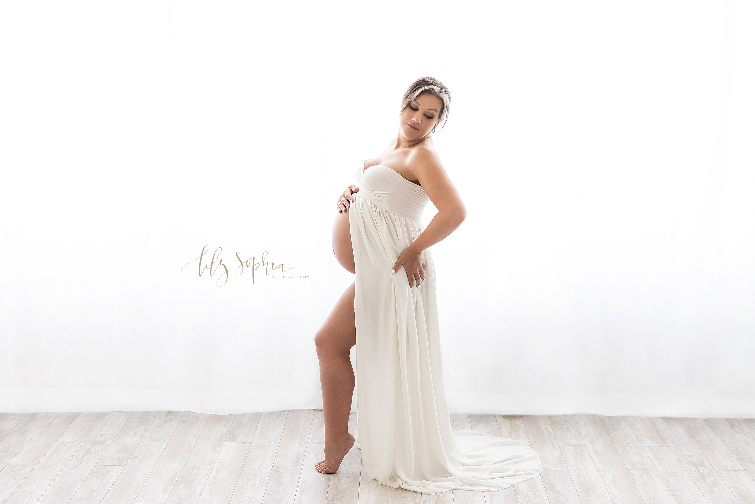  Profile maternity portrait of a pregnant mother standing in a natural light studio wearing a strapless split front knit gown as she places her right hand on top of her bare belly and her left hand on her left hip while looking over her left shoulder