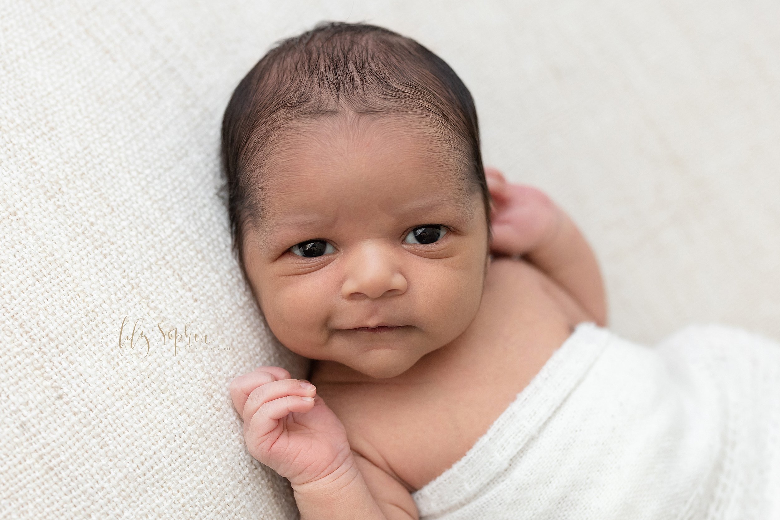  Newborn portrait of an Indian infant boy as he lies on his back wide awake and smirks taken near Alpharetta in Atlanta in natural light in a photography studio. 