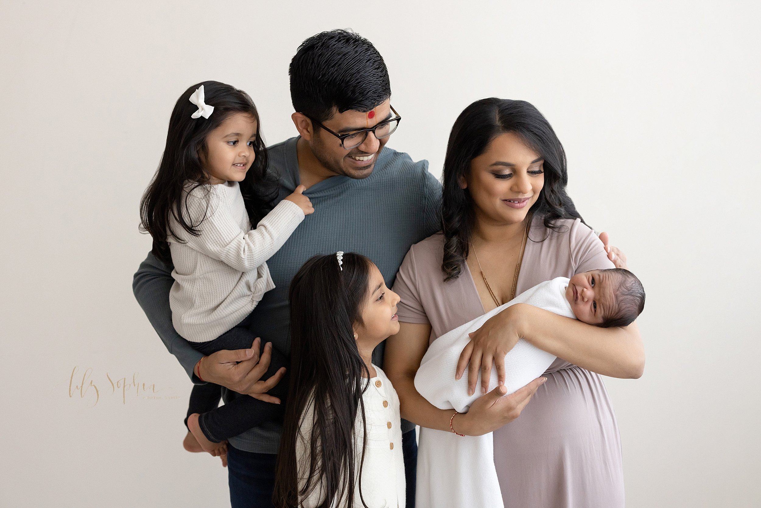  Family newborn photo of an Indian mother holding her newborn baby boy in her arms  with her husband standing behind her holding their toddler daughter in his right arm and their young daughter standing in front of dad as they all admire the baby tak