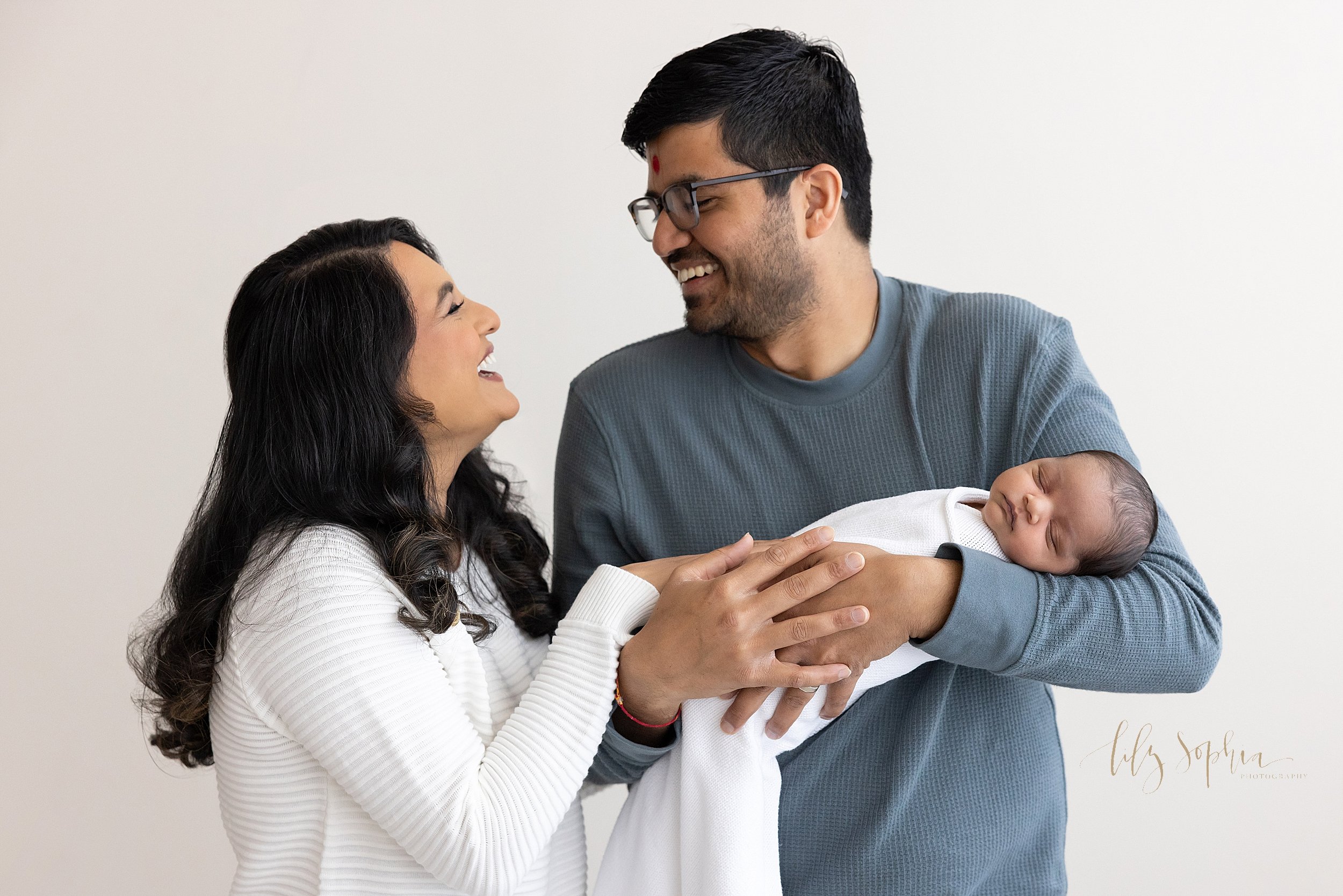  An Indian father cradles his son in his arms as he looks over his right shoulder to look into his wife’s eyes as the two of them laugh taken near Oakhurst in Atlanta in a studio using natural light. 