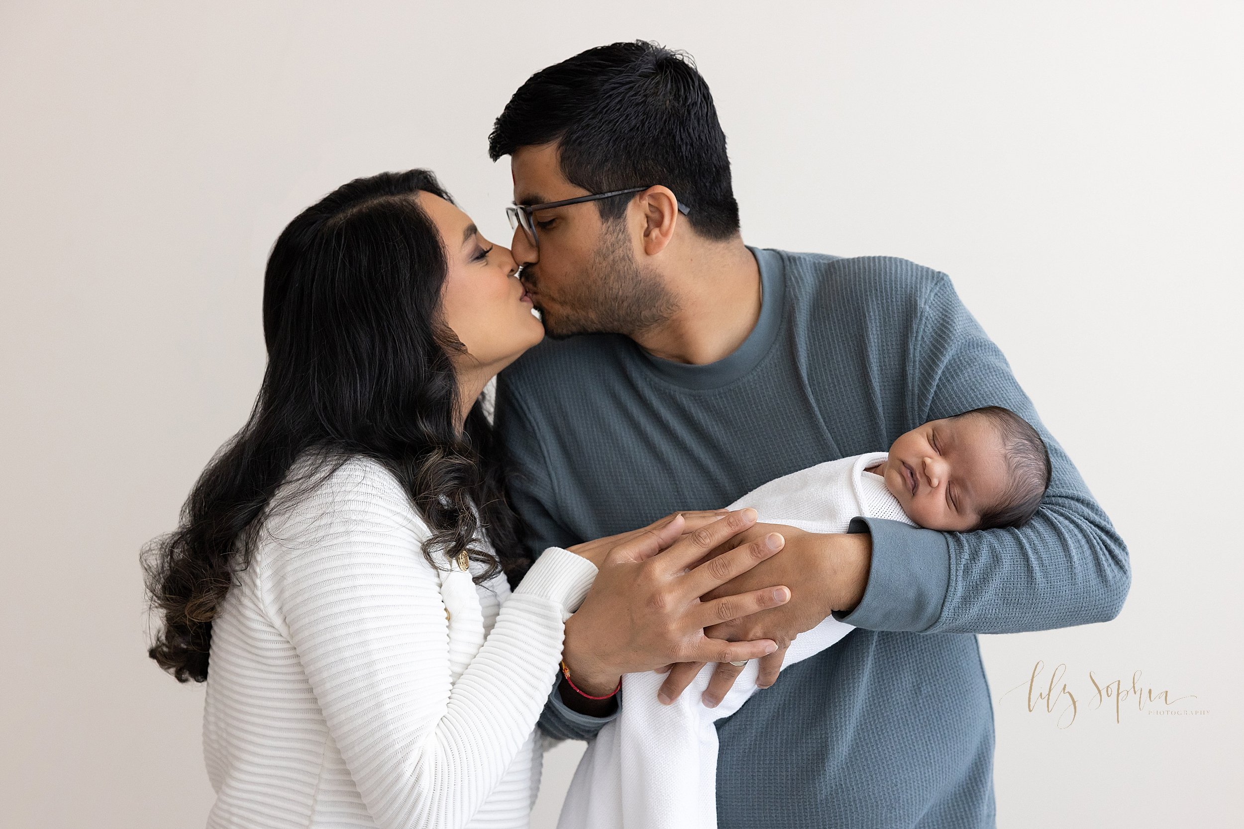  Newborn photo session of an Indian father cradling his newborn son in his arms as his wife stands to his right side and the two of them kiss taken near Smyrna in Atlanta in a studio using natural light. 