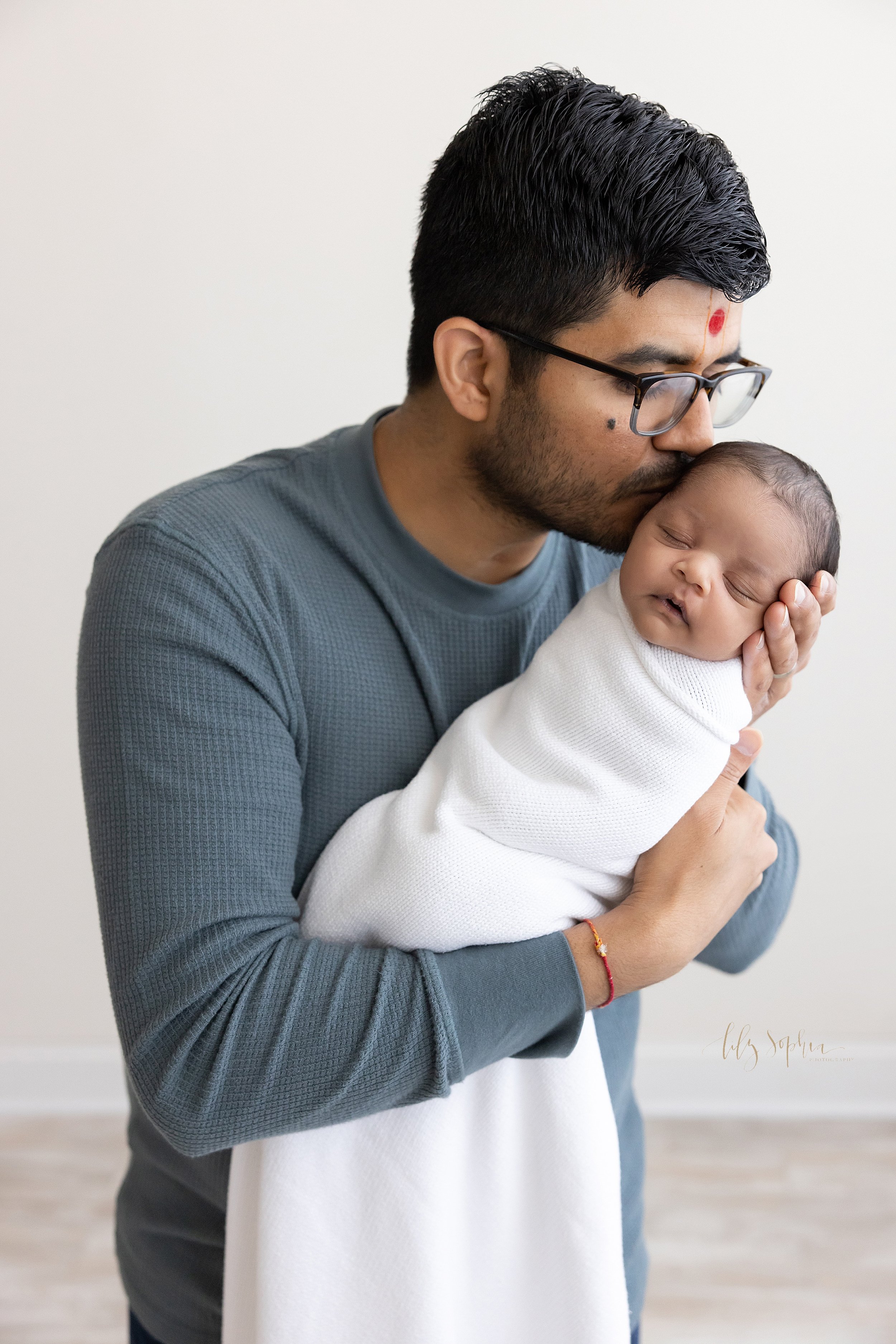  An Indian father holds his newborn son in his arms as he kisses his son’s head while the infant peacefully sleeps taken near Roswell in Atlanta in a natural light studio. 