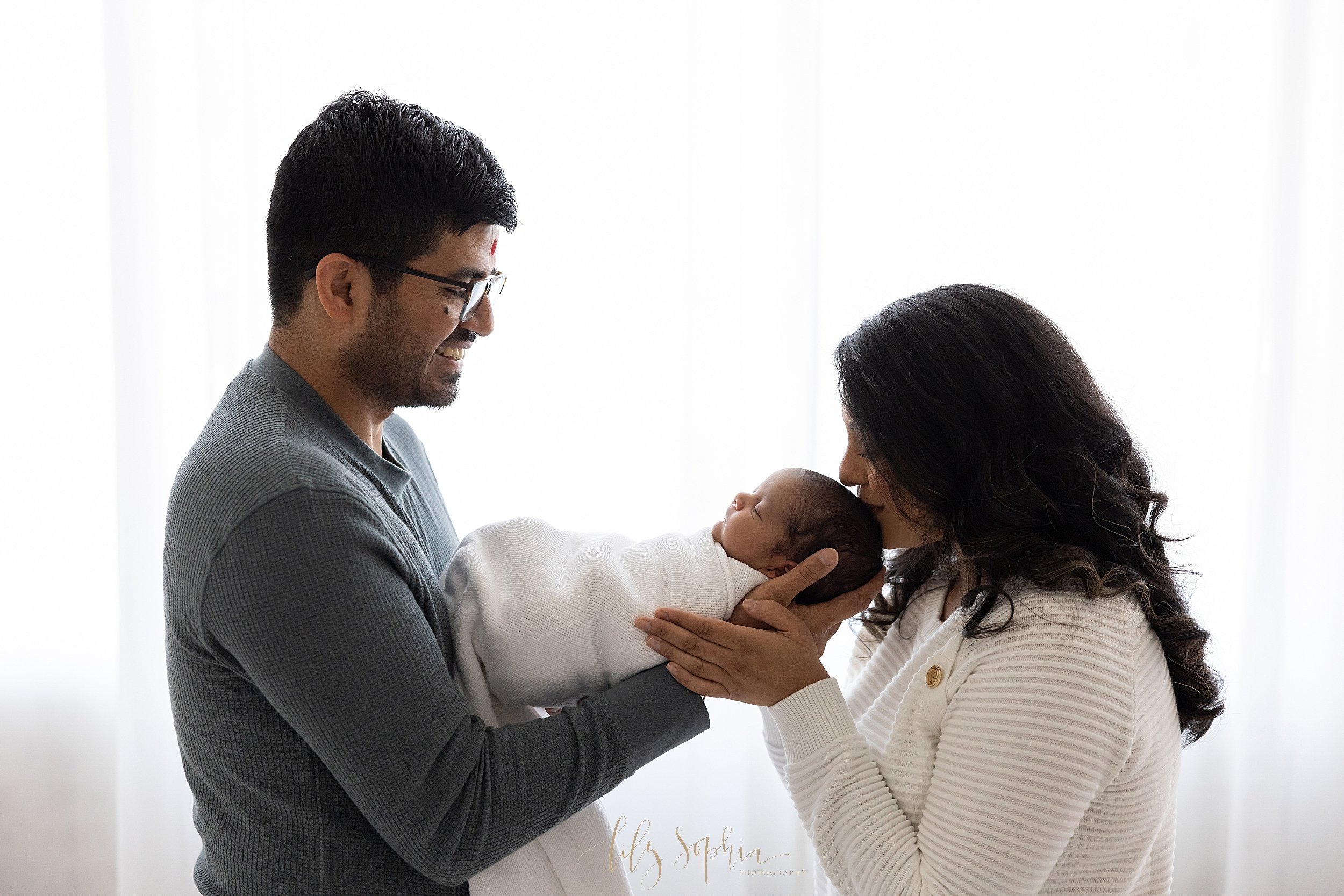  Newborn portrait of an Indian father holding his son in his hands in front of him while his wife faces him, places her hands on his and kisses the crown of her infant’s son’s head as the couple stand in front of a window streaming natural light in a