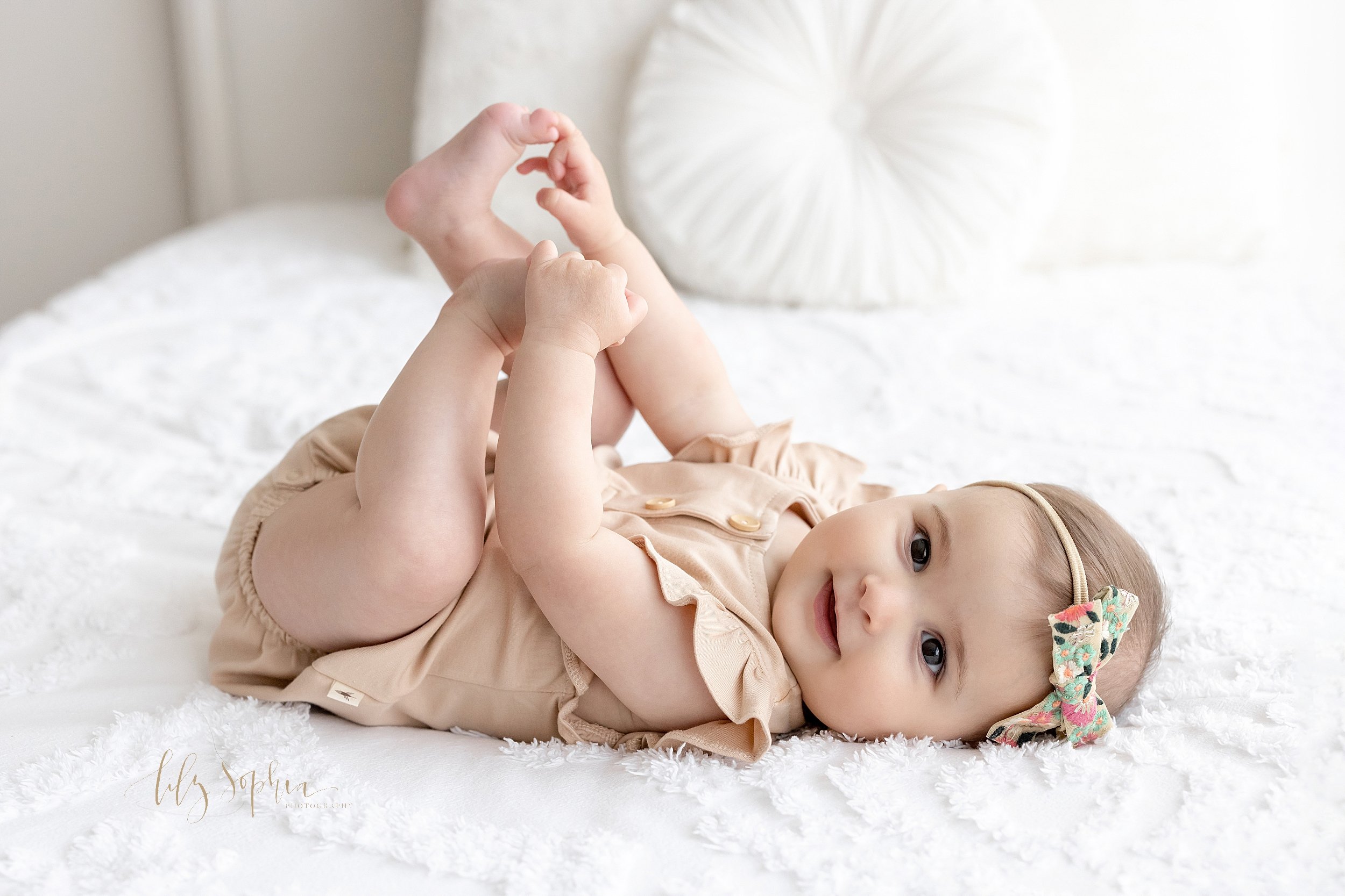  First birthday photo session of a one year old little girl as she lies on her back on a bed in a natural light studio and plays with her toes taken near Midtown in Atlanta, Georgia. 